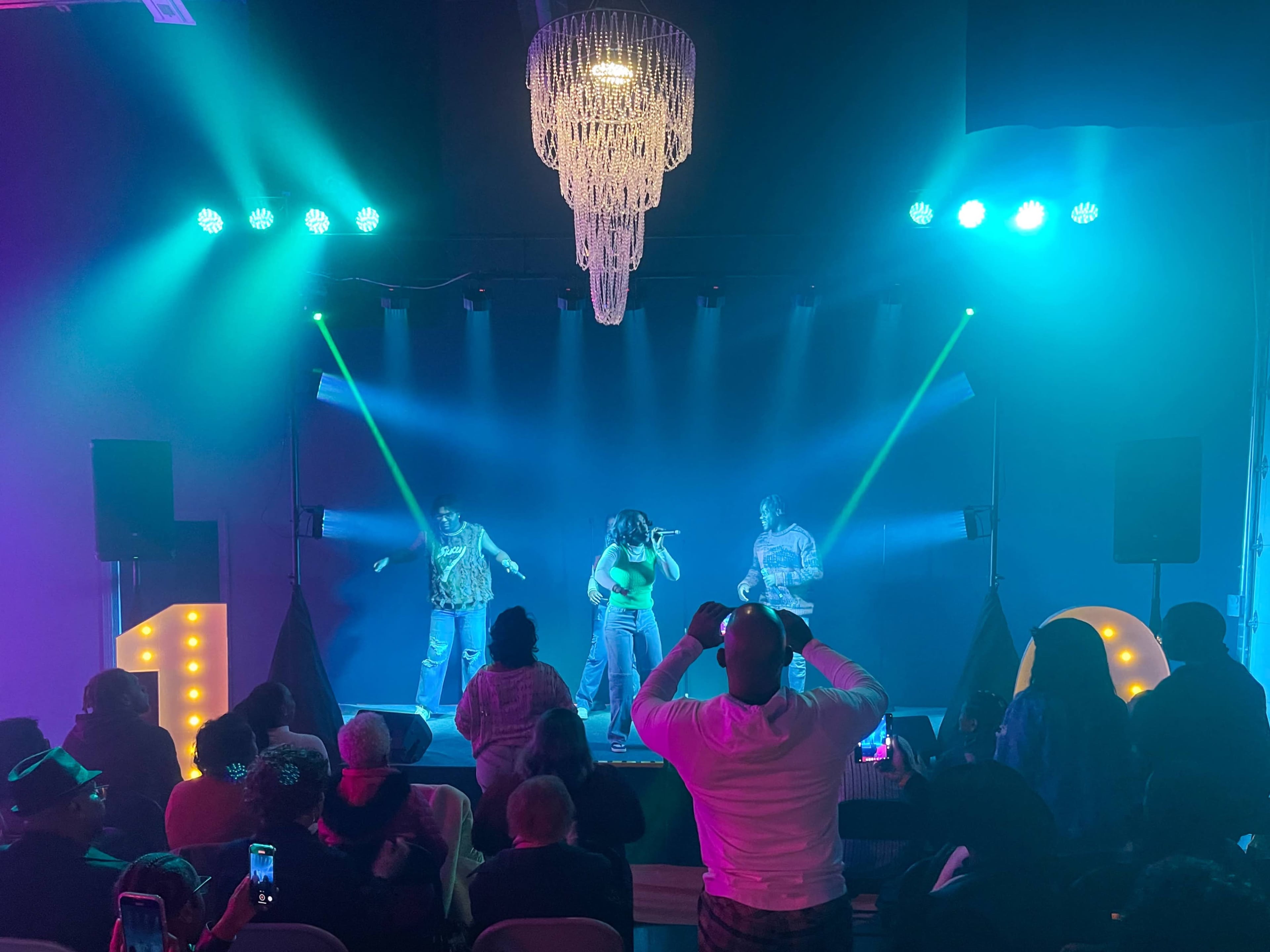 A live performance takes place on stage, illuminated by colorful lights, with an audience seated in front.