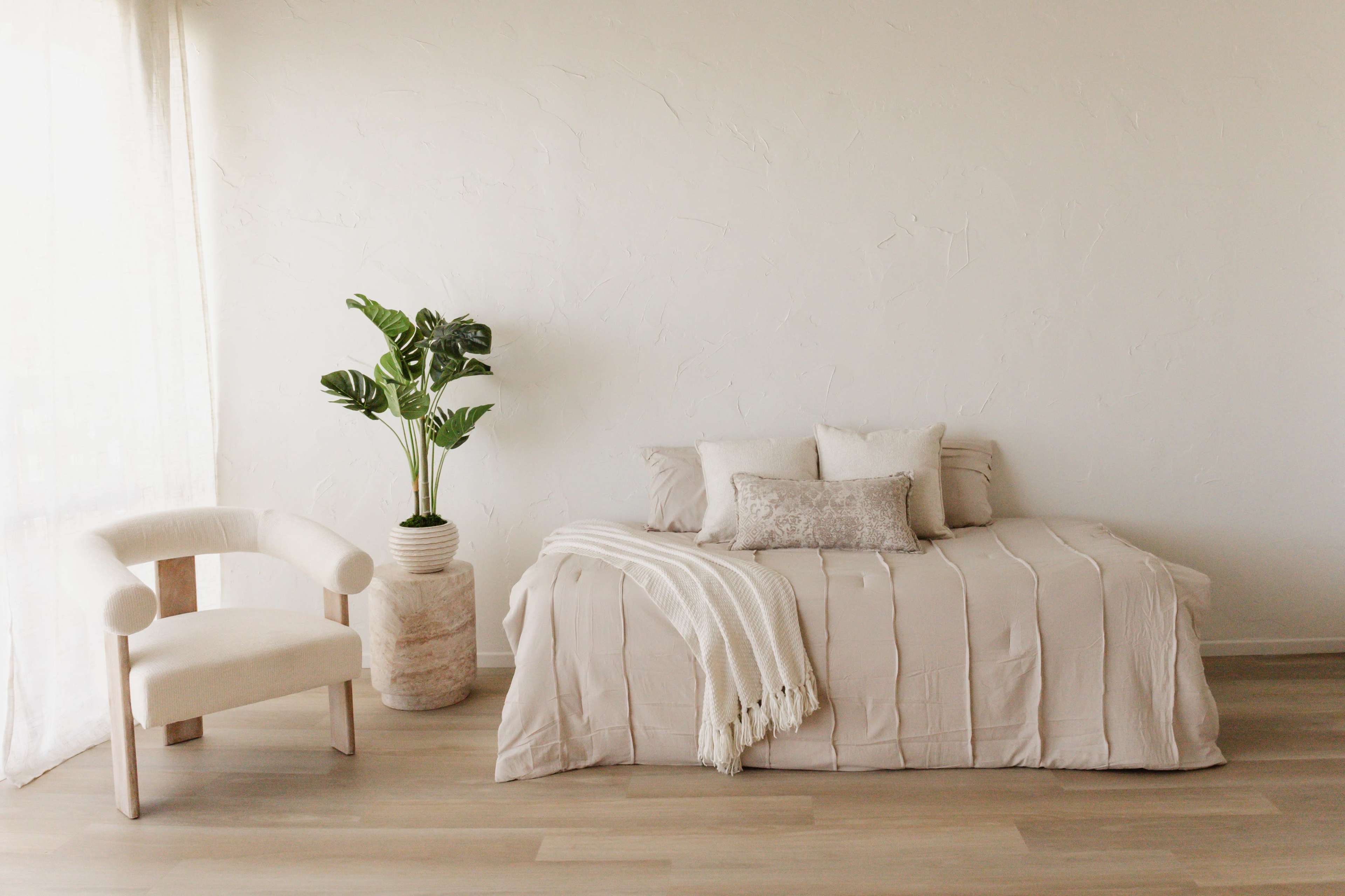 The room features a light-colored bed with decorative pillows and a chair beside a potted plant against a textured white wall.