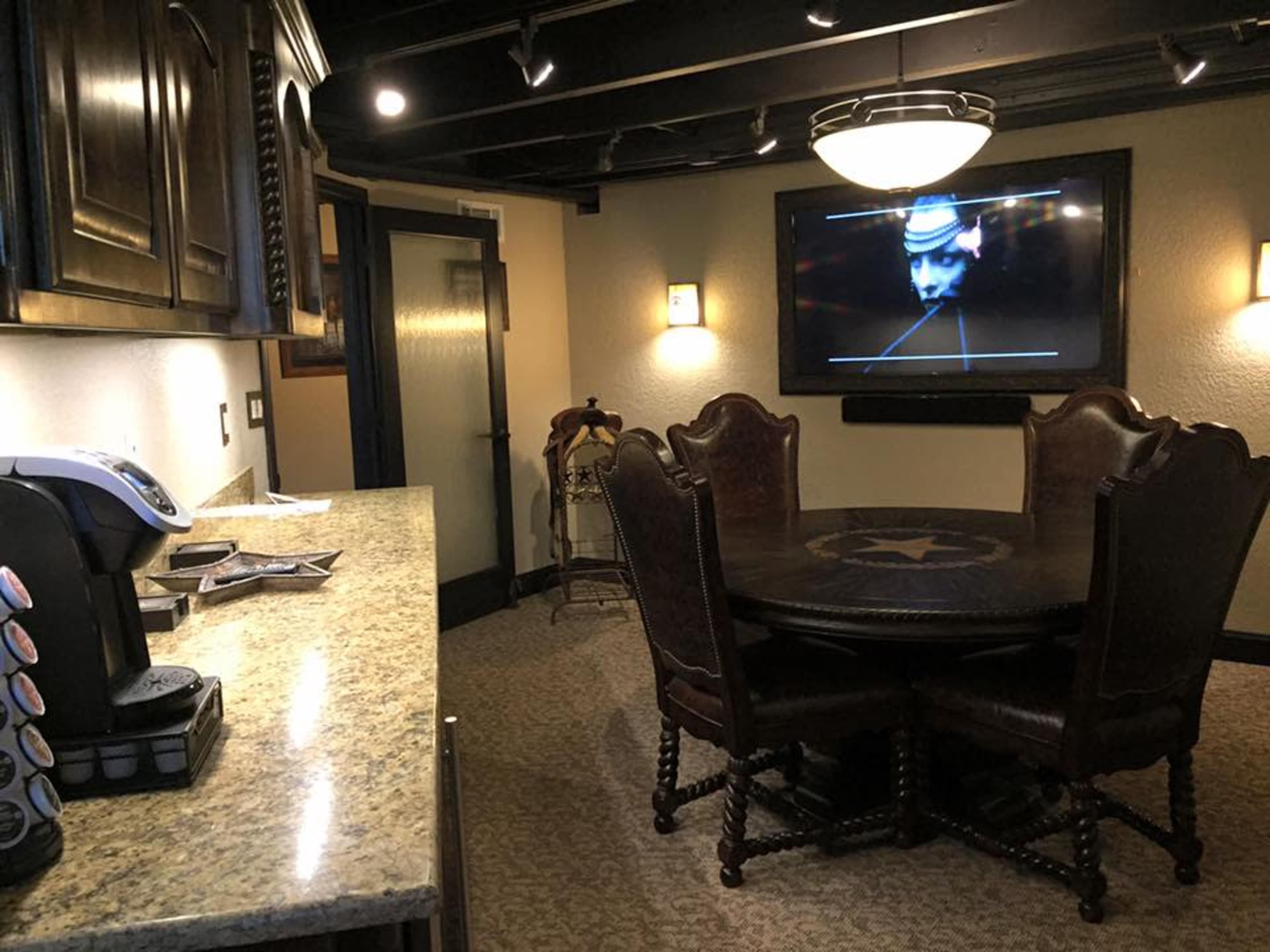 The image shows a kitchen area with a granite countertop, a coffee maker, and a round dining table surrounded by chairs, with a large television mounted on the wall displaying a dark image.