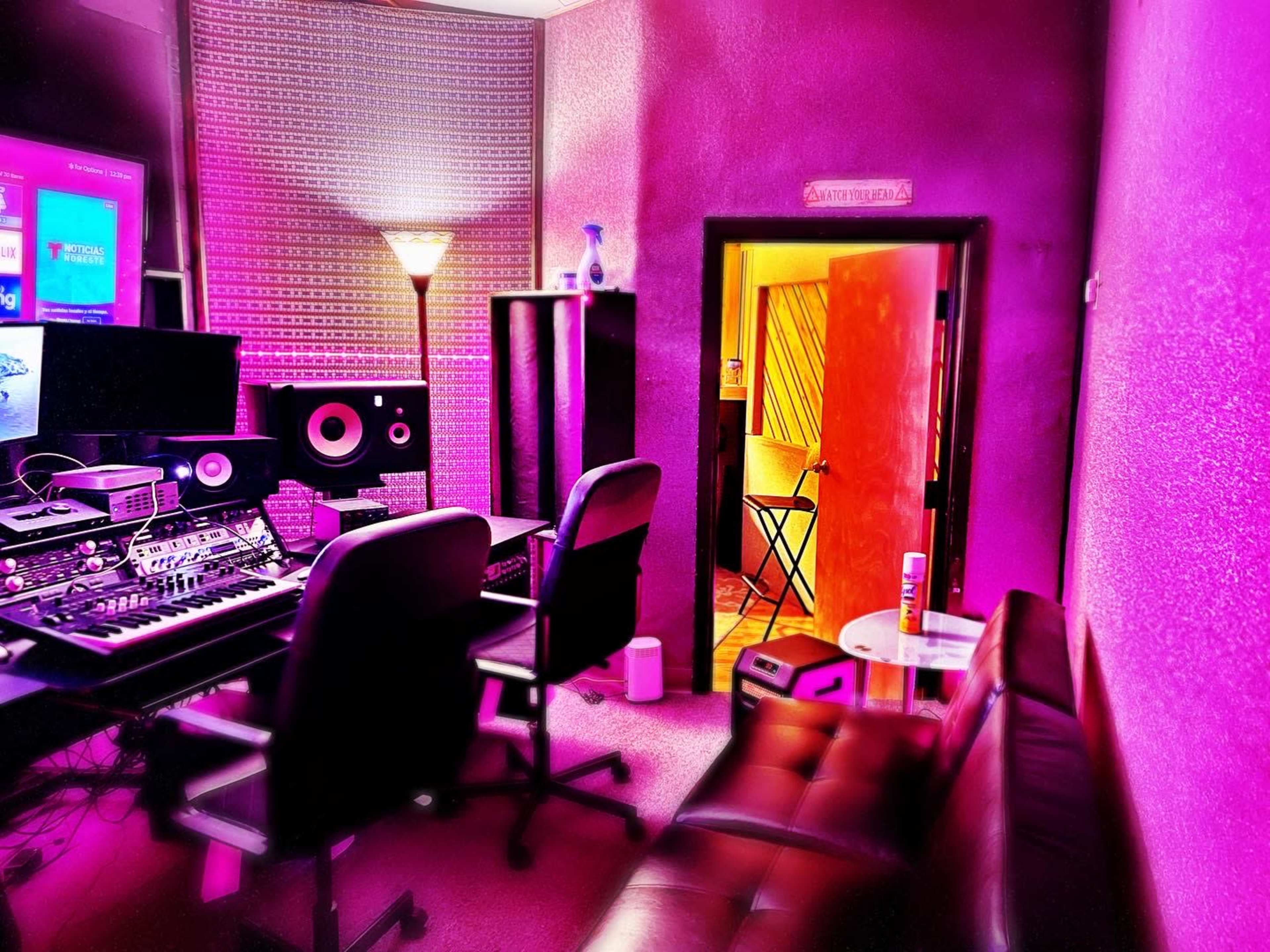 The image shows a music production studio with two black chairs, a mixing console, and a doorway leading to a recording booth.