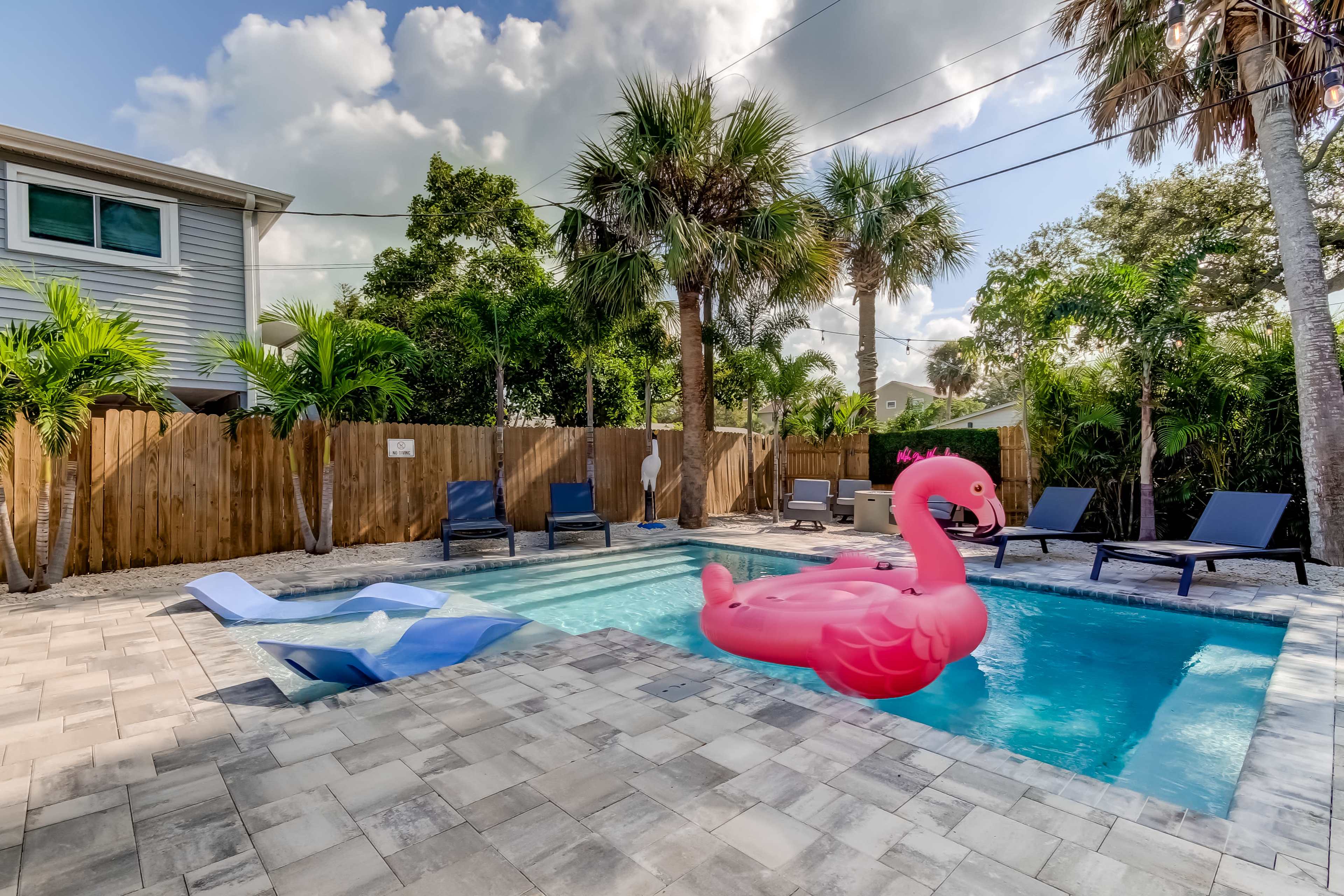 A backyard pool area featuring a pink flamingo float, lounge chairs, palm trees, and a fenced perimeter.