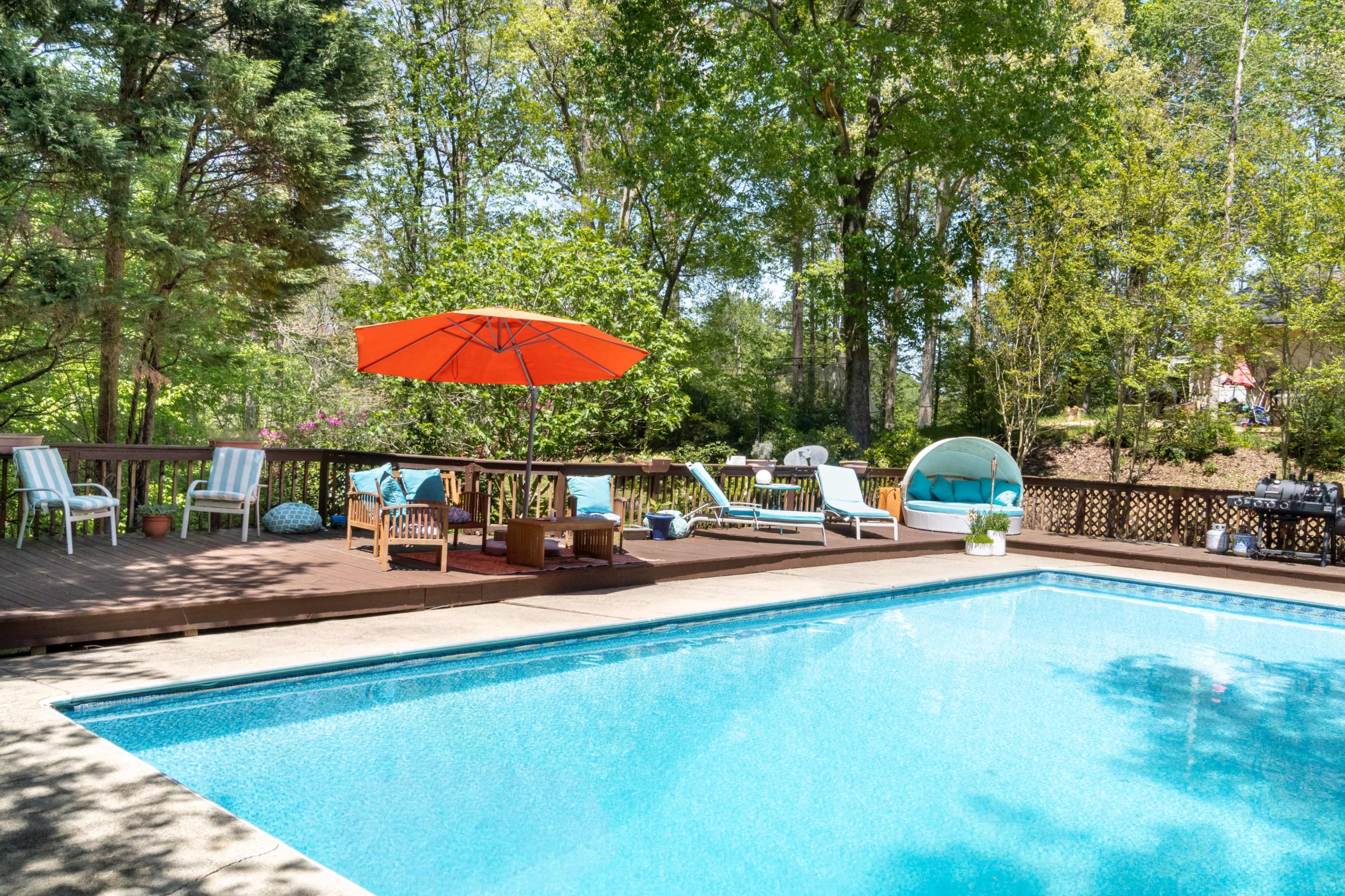 A backyard features a swimming pool surrounded by lounge chairs, a table with an orange umbrella, and a shaded seating area amidst trees and greenery.