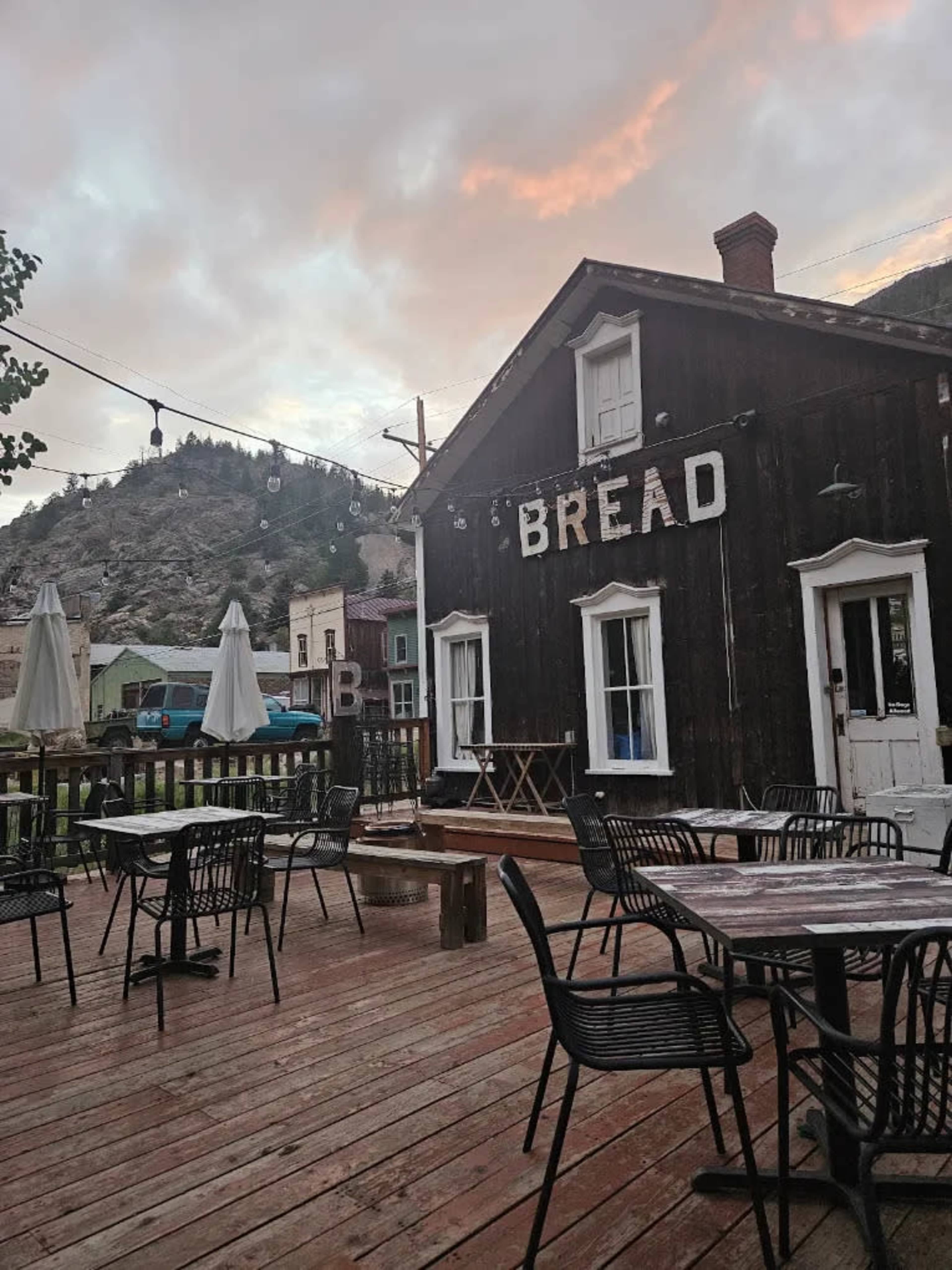 The image shows a rustic wooden building with the word "BREAD" painted on its side, surrounded by outdoor dining furniture on a deck with a mountainous backdrop.
