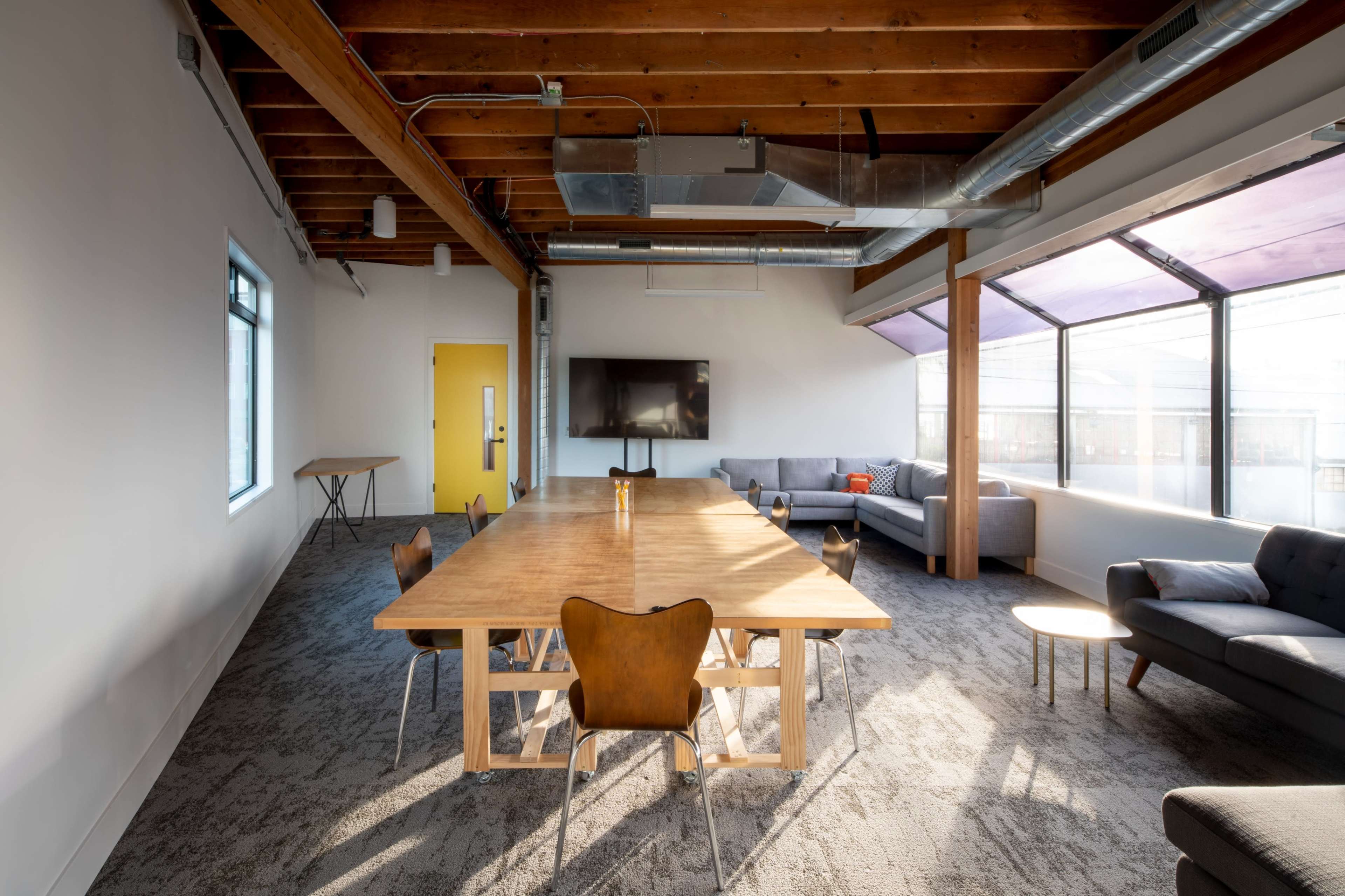 A spacious conference room features a large wooden table surrounded by chairs, with a seating area and large windows allowing natural light.