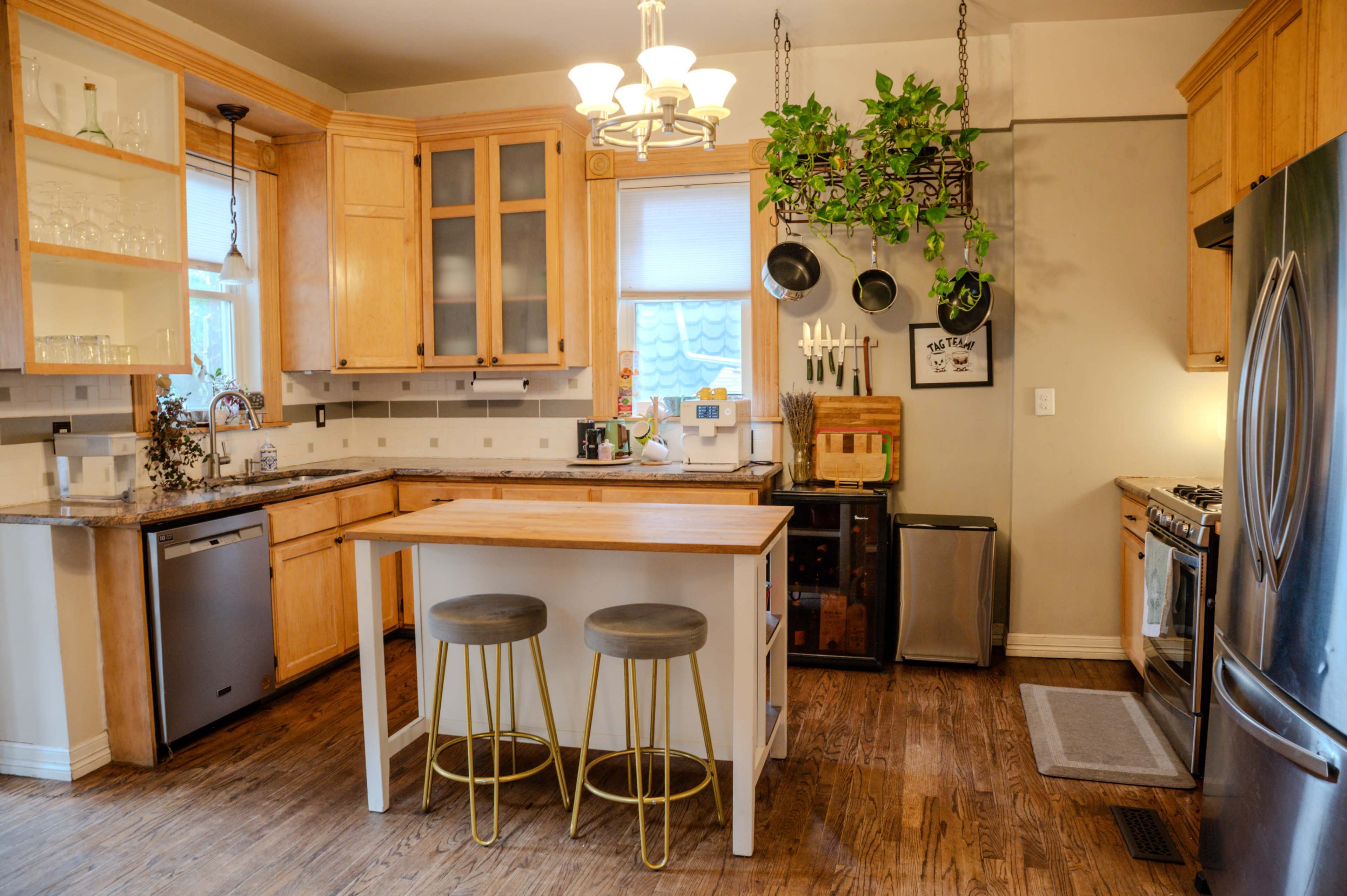 The image shows a modern kitchen featuring wooden cabinets, a central island with two stools, and stainless steel appliances, along with plants hanging from the ceiling.