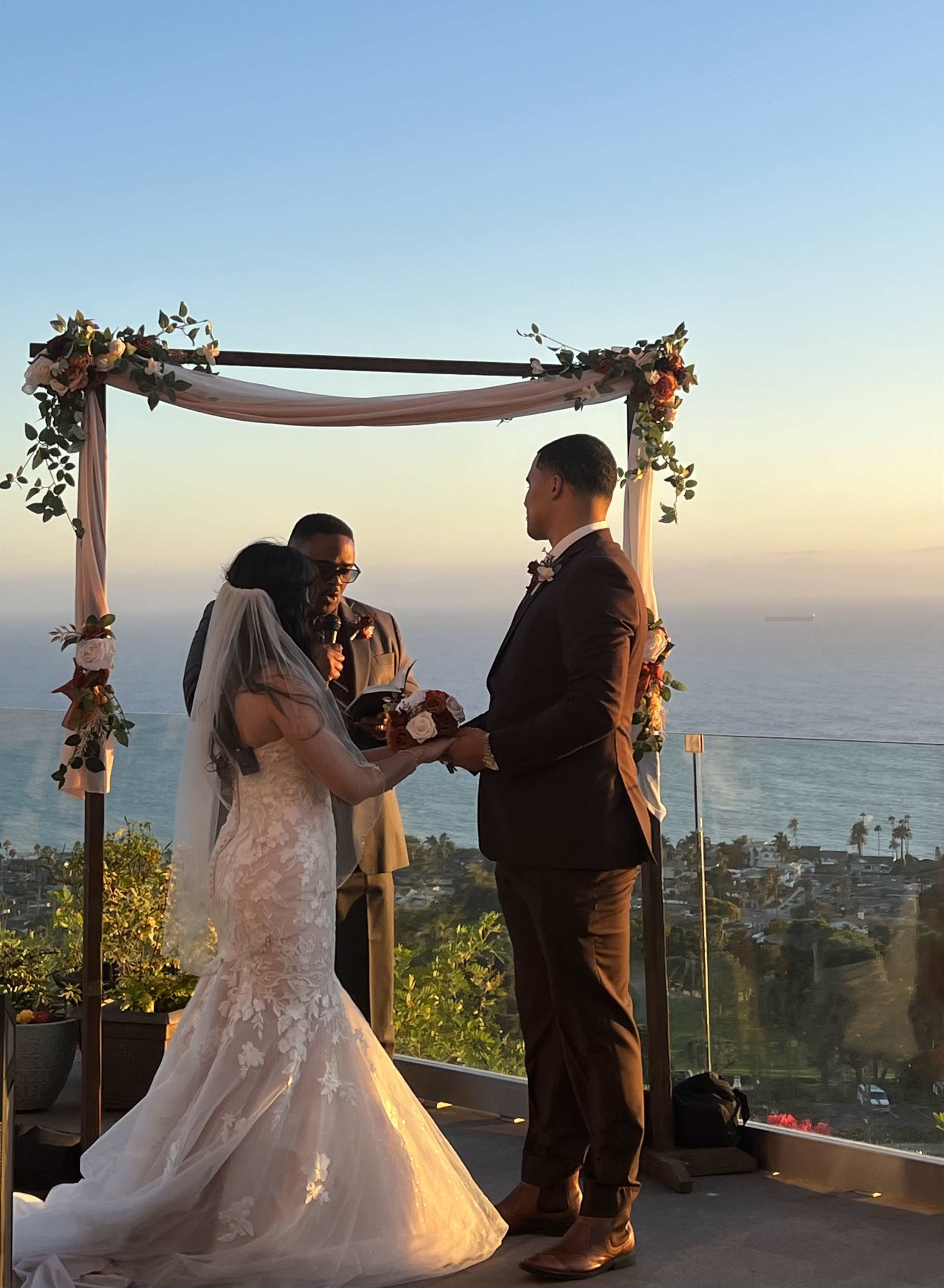 A couple stands under a floral arch during their wedding ceremony with a scenic ocean view in the background.
