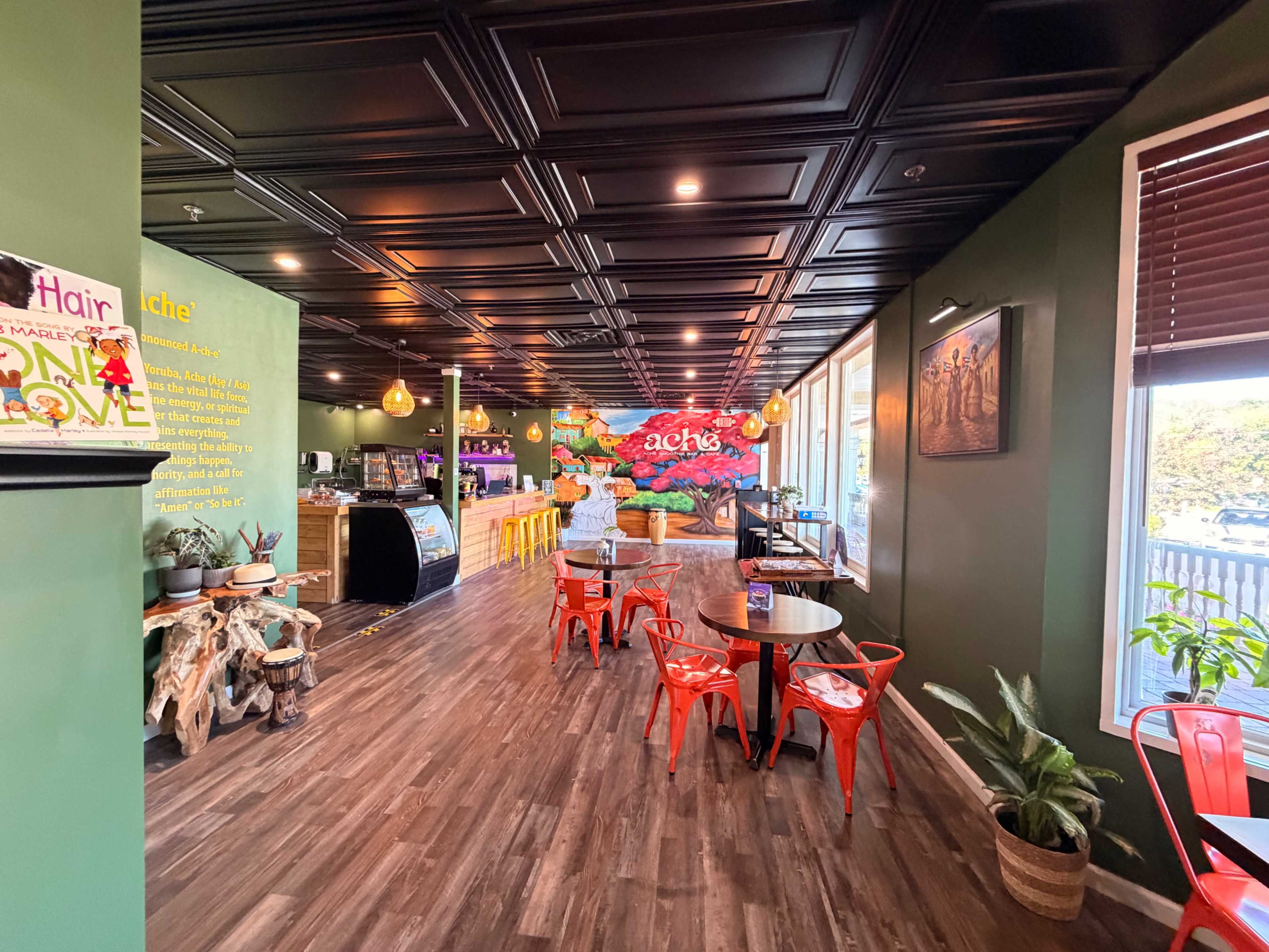 The image shows an interior of a colorful café featuring wooden flooring, green walls, and red chairs arranged around small tables, with a mural on one wall and a counter area in the background.