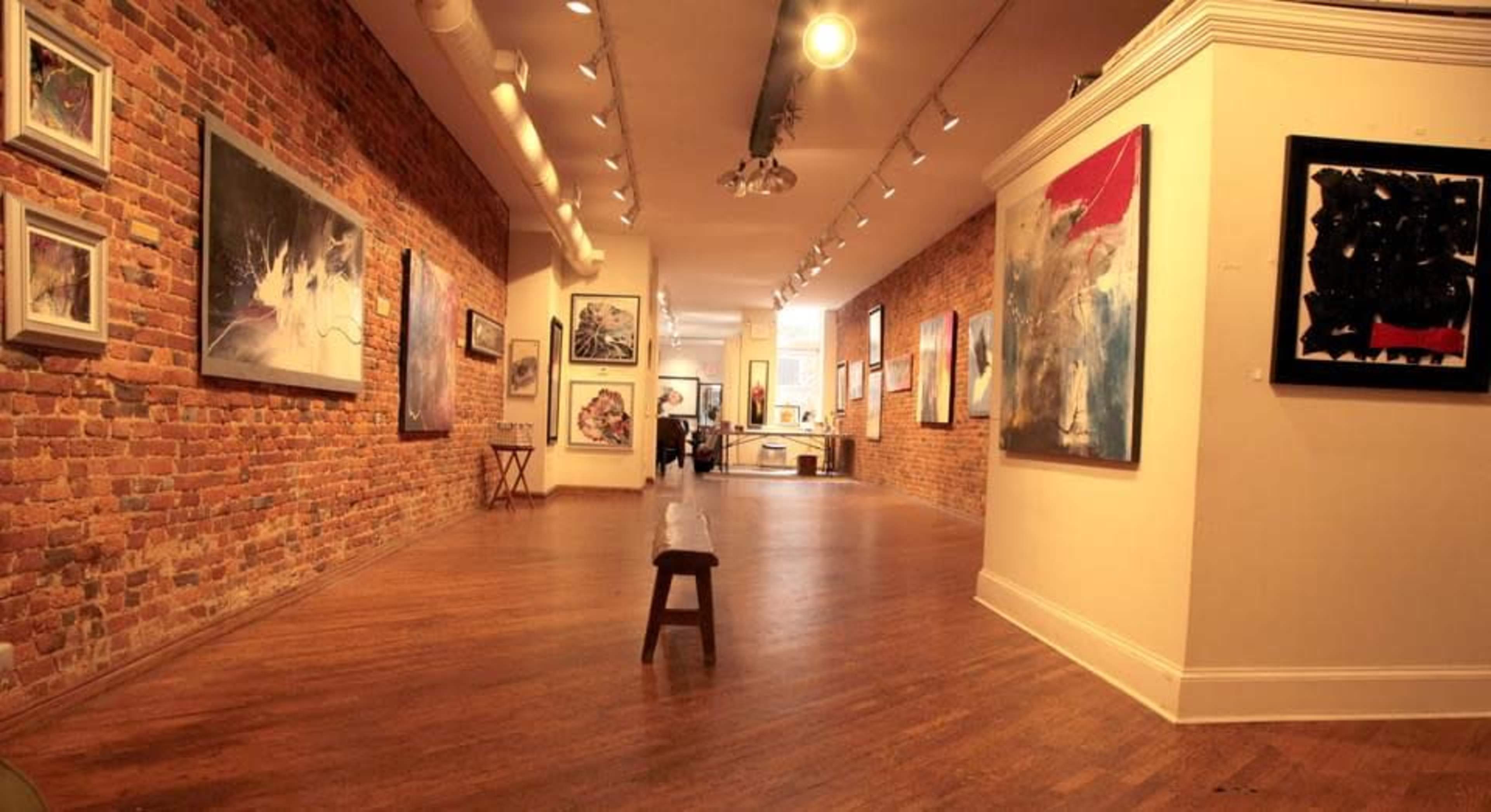 The interior of an art gallery with exposed brick walls and a variety of paintings displayed on the walls, along with wooden flooring and a bench in the foreground.