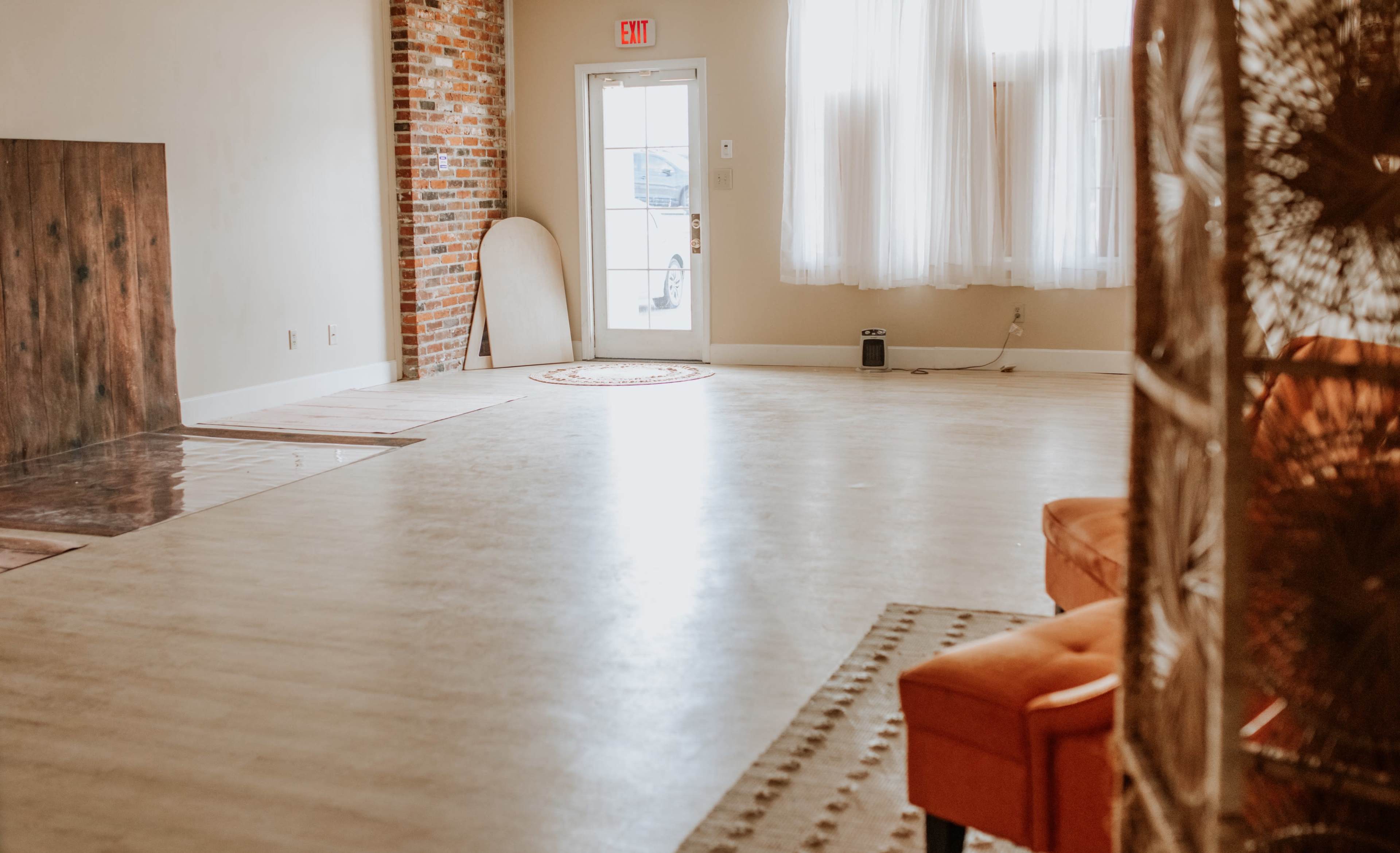 The image shows a spacious, empty room with a large window, exposed brick wall, and a few pieces of furniture, including an orange couch.