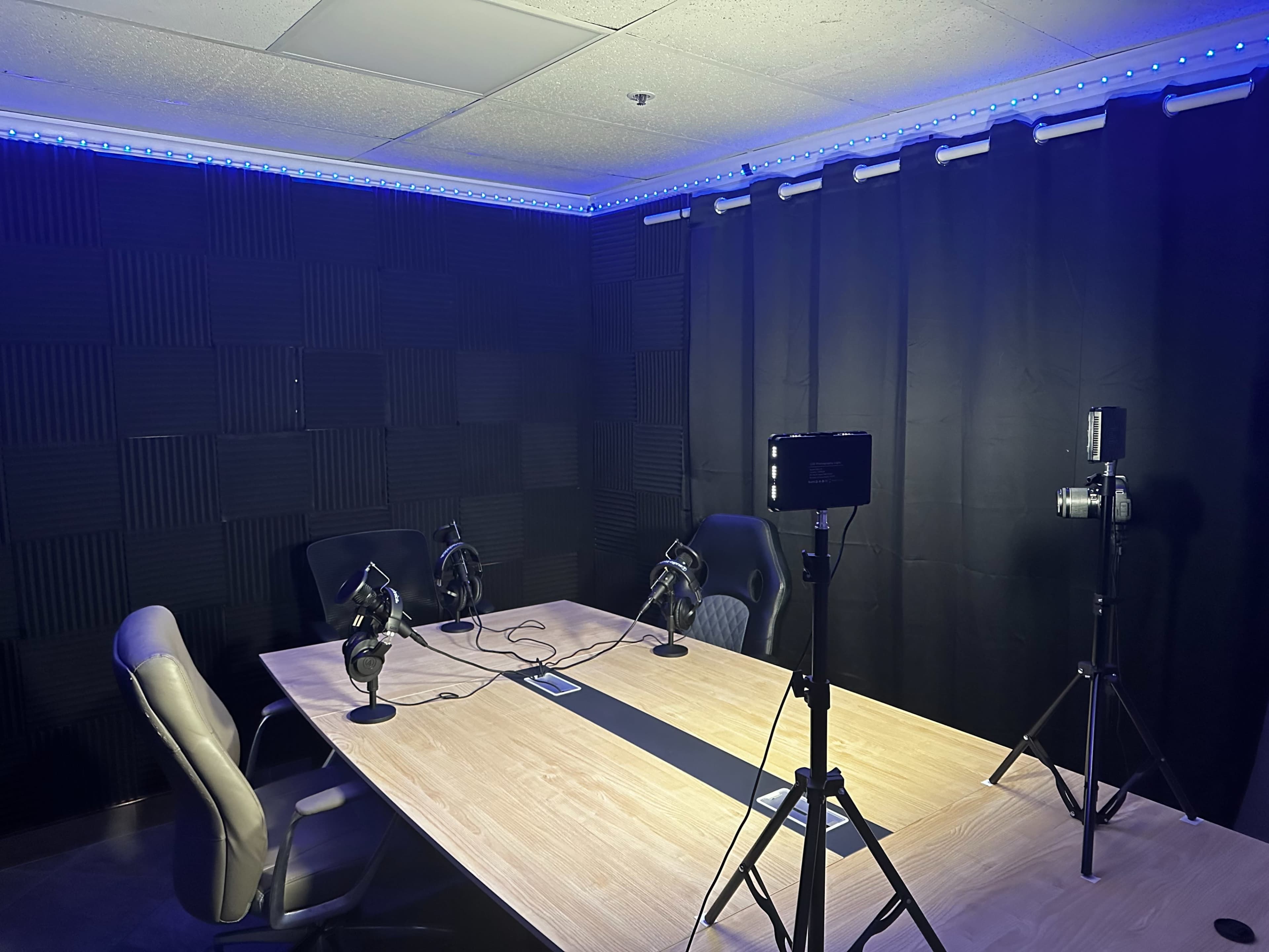 A recording studio featuring a large wooden table with several microphones, chairs, and lighting equipment, surrounded by sound-absorbing wall panels.