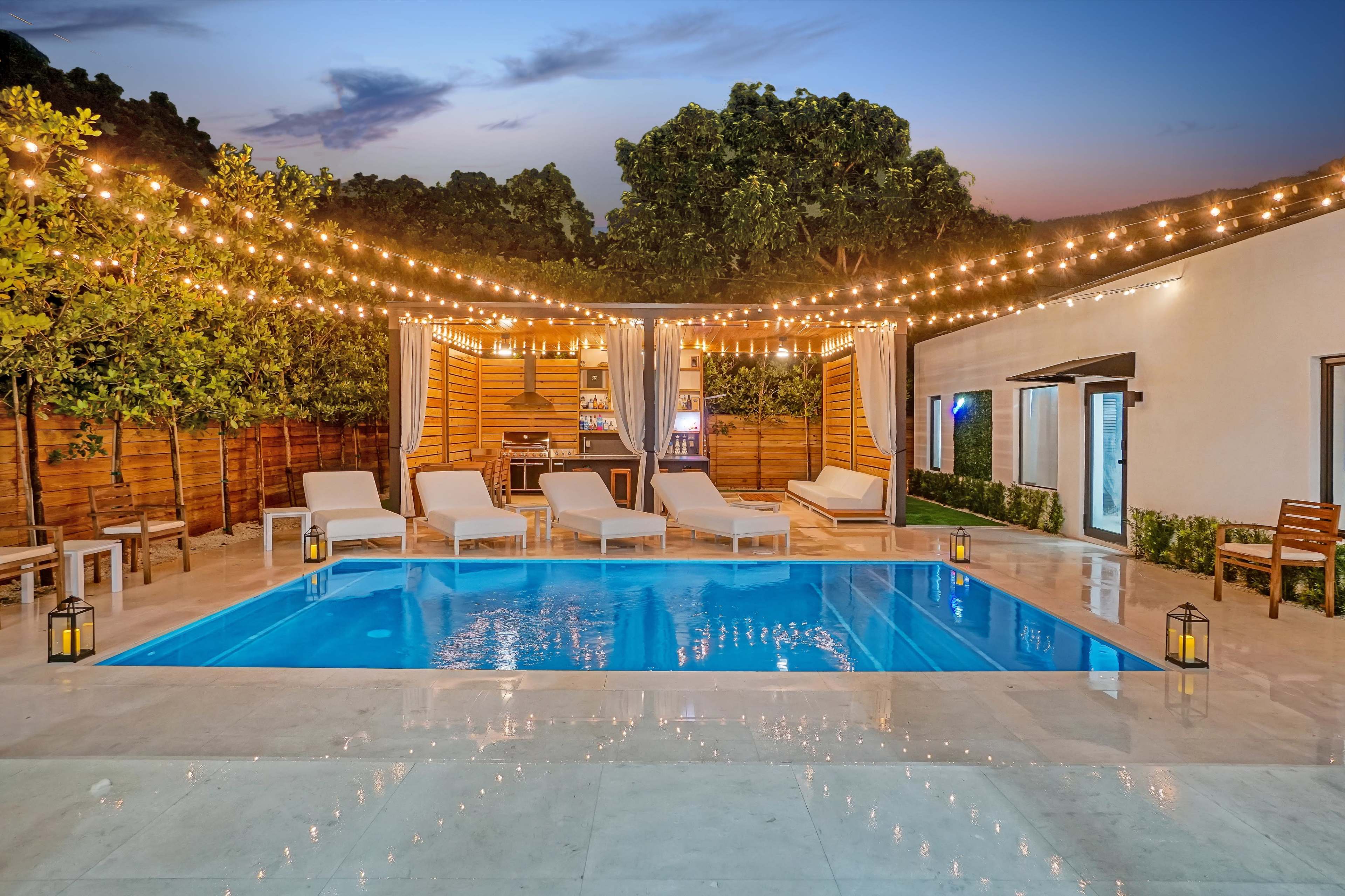 The image features a stylish outdoor pool area illuminated by string lights, with comfortable loungers surrounding the water and lush greenery in the background.