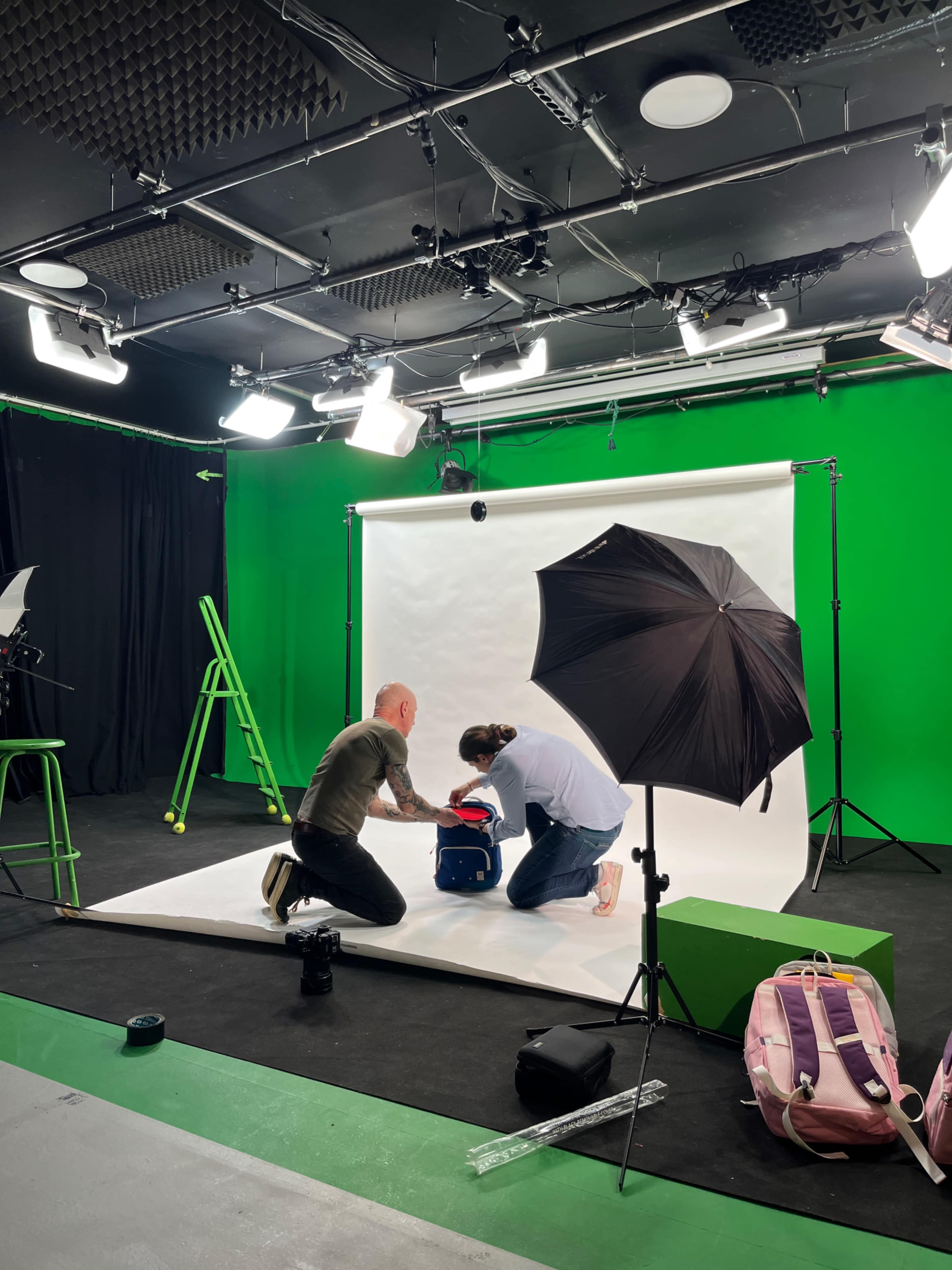 A photographer is setting up a shot with a model and a backpack in a studio with green walls and various lighting equipment.