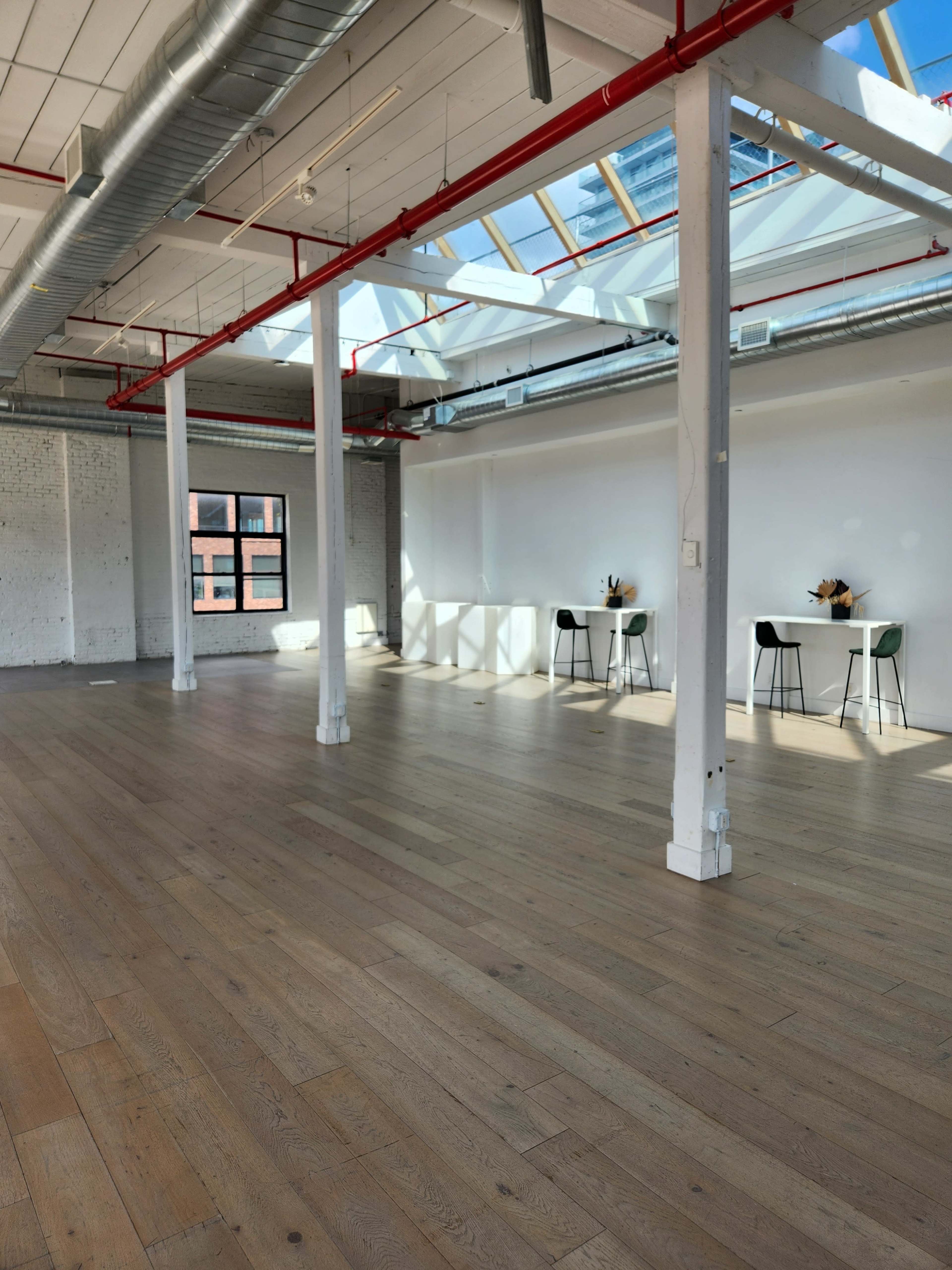 The image shows a spacious, well-lit room with wooden flooring, large windows, and a few tables with chairs near the walls.