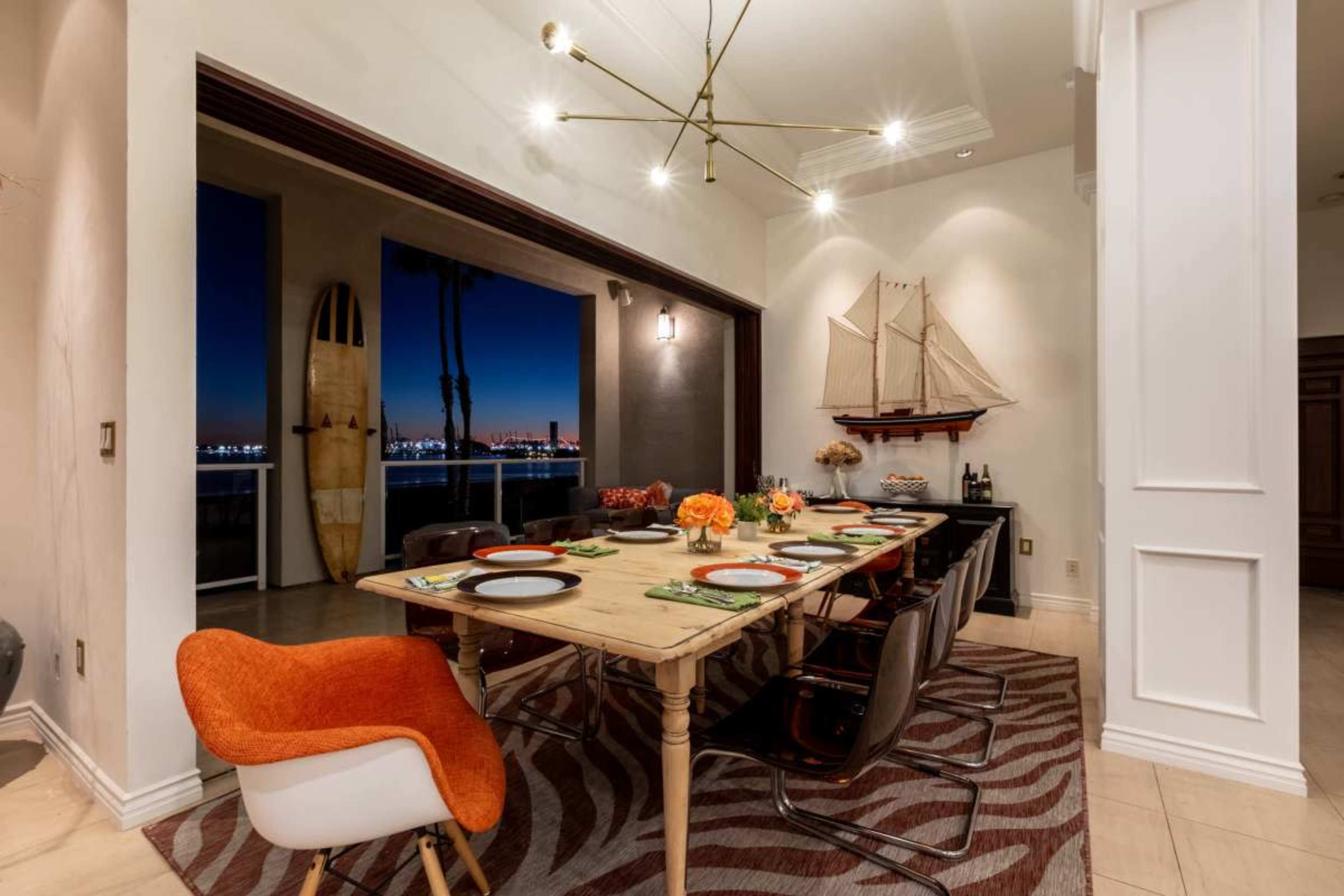 A dining room features a large wooden table set for a meal, with a view of palm trees and the city skyline visible through sliding glass doors.