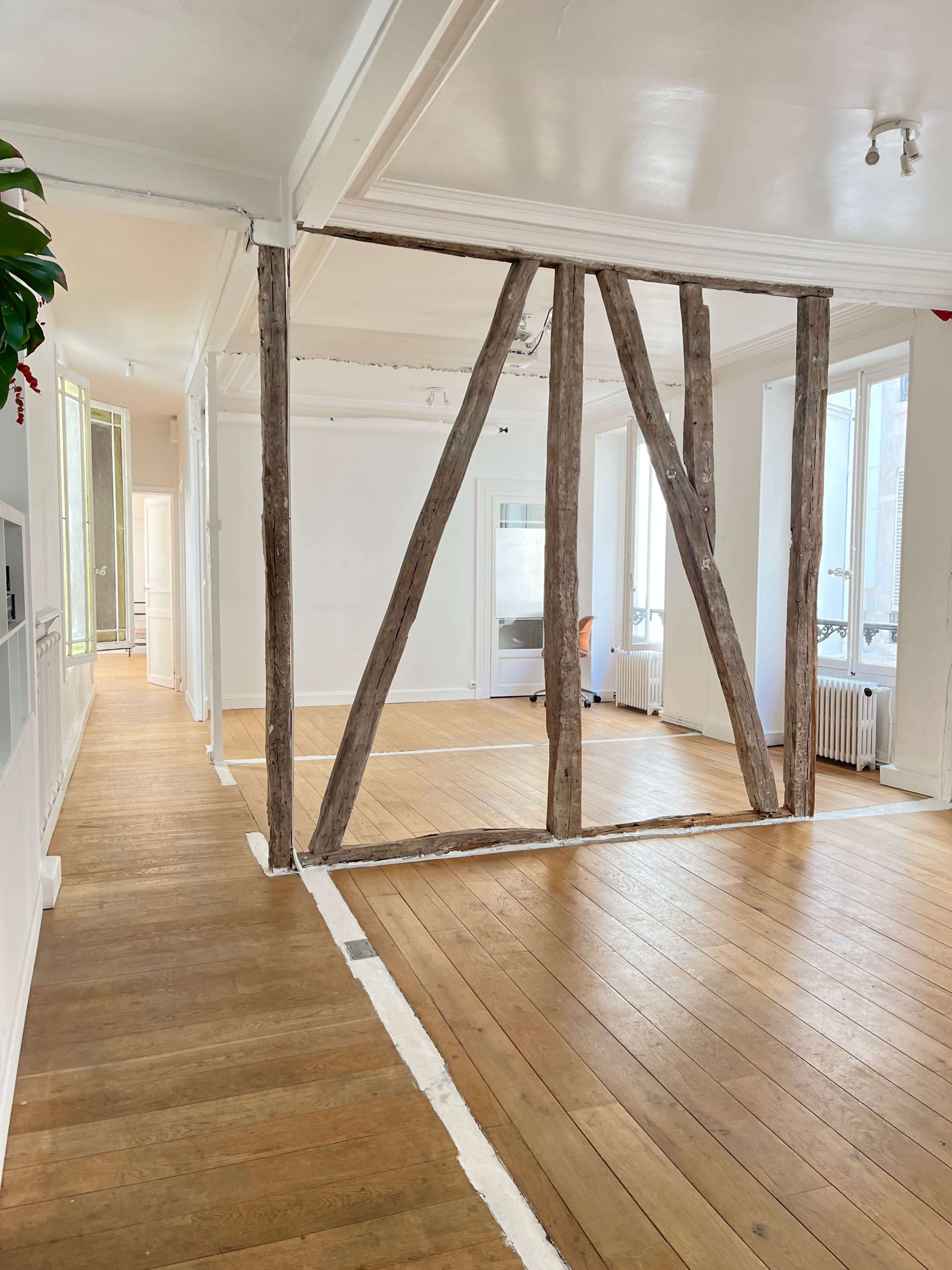 The image shows a spacious interior with wooden beams dividing an open layout, featuring large windows and a wooden floor.