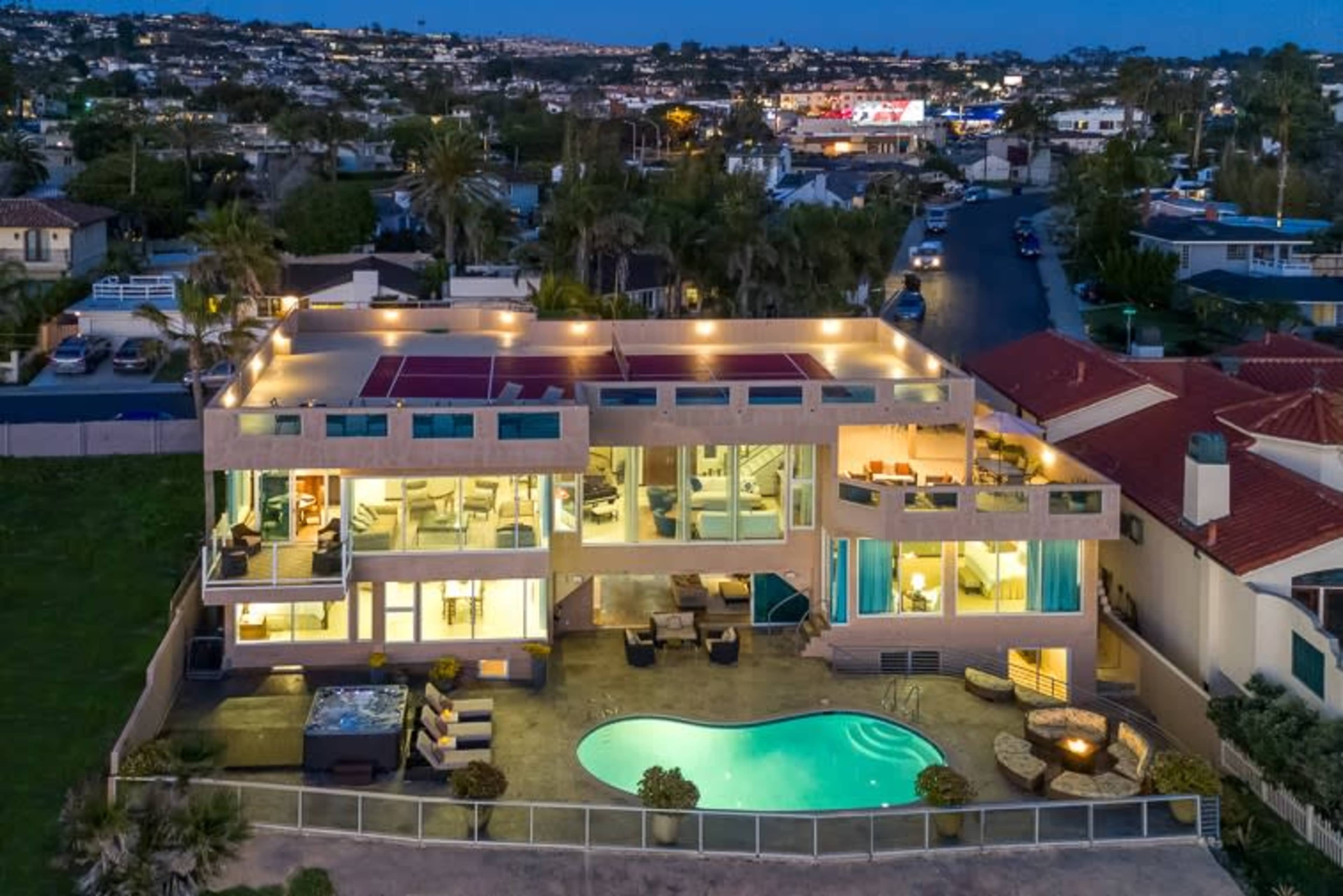 A modern multi-level house with large glass windows is situated beside a swimming pool, surrounded by palm trees and overlooking a suburban neighborhood at dusk.