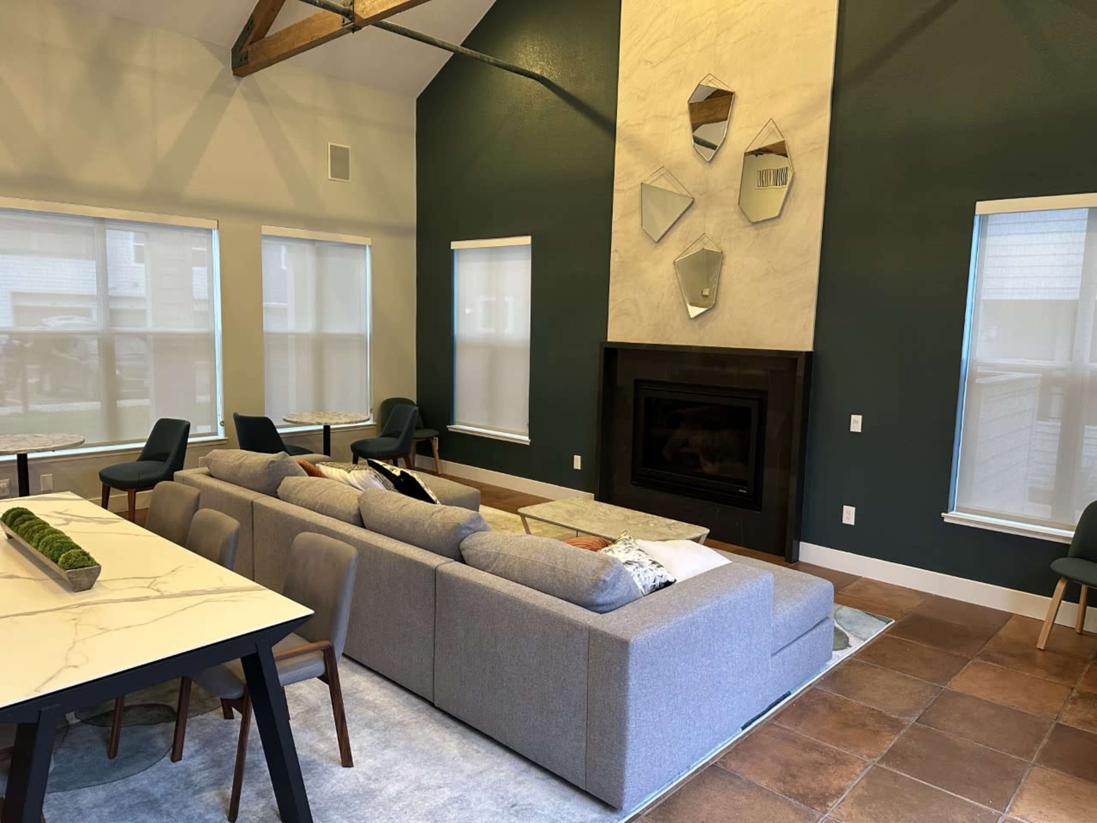 A modern living room features a gray sectional sofa, a marble dining table, and decorative mirrors above a black fireplace, with large windows allowing natural light.