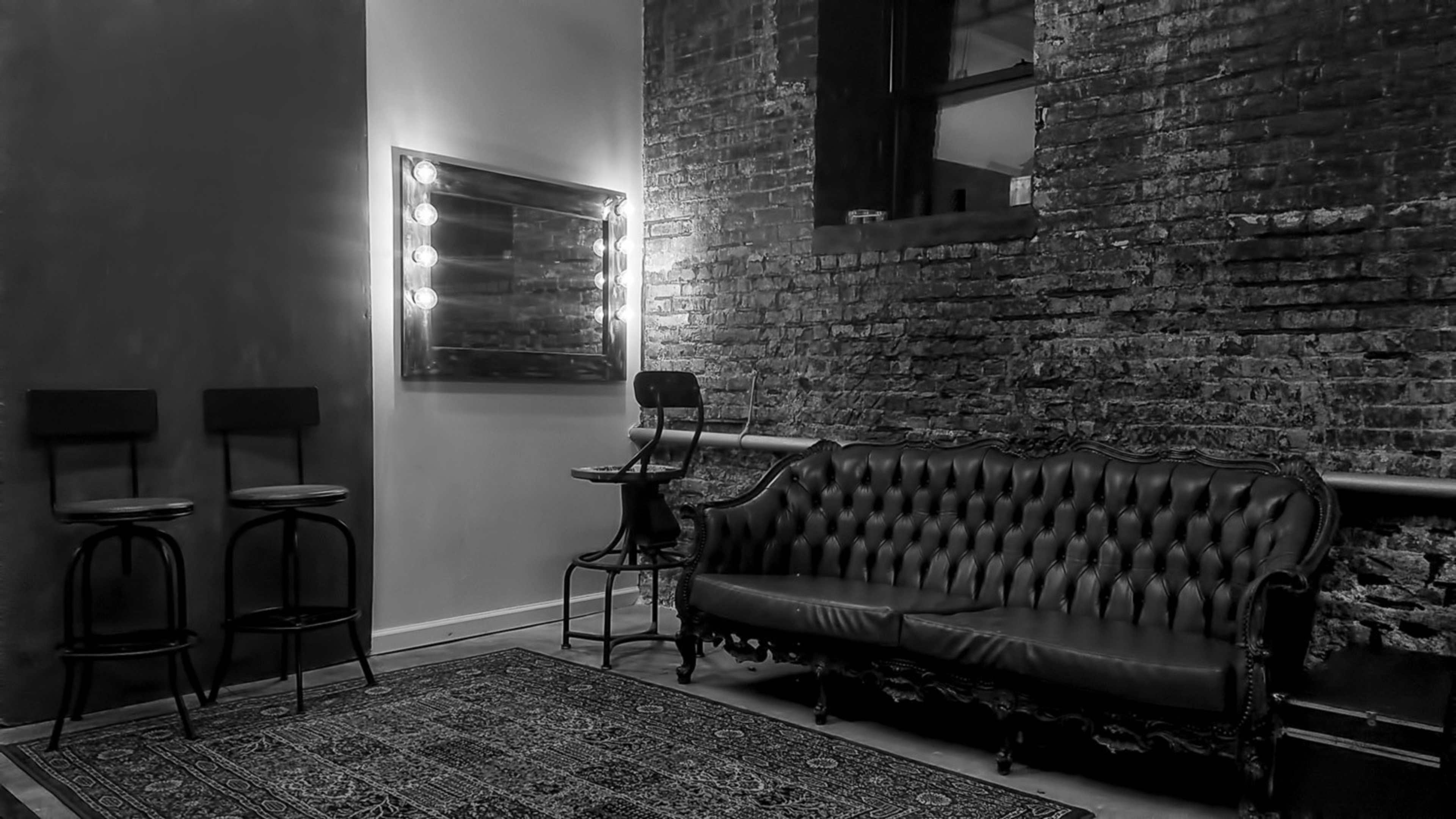 The image shows a rustic room with a tufted leather sofa, a small round table, and tall bar stools against a brick wall, illuminated by a lighted mirror.