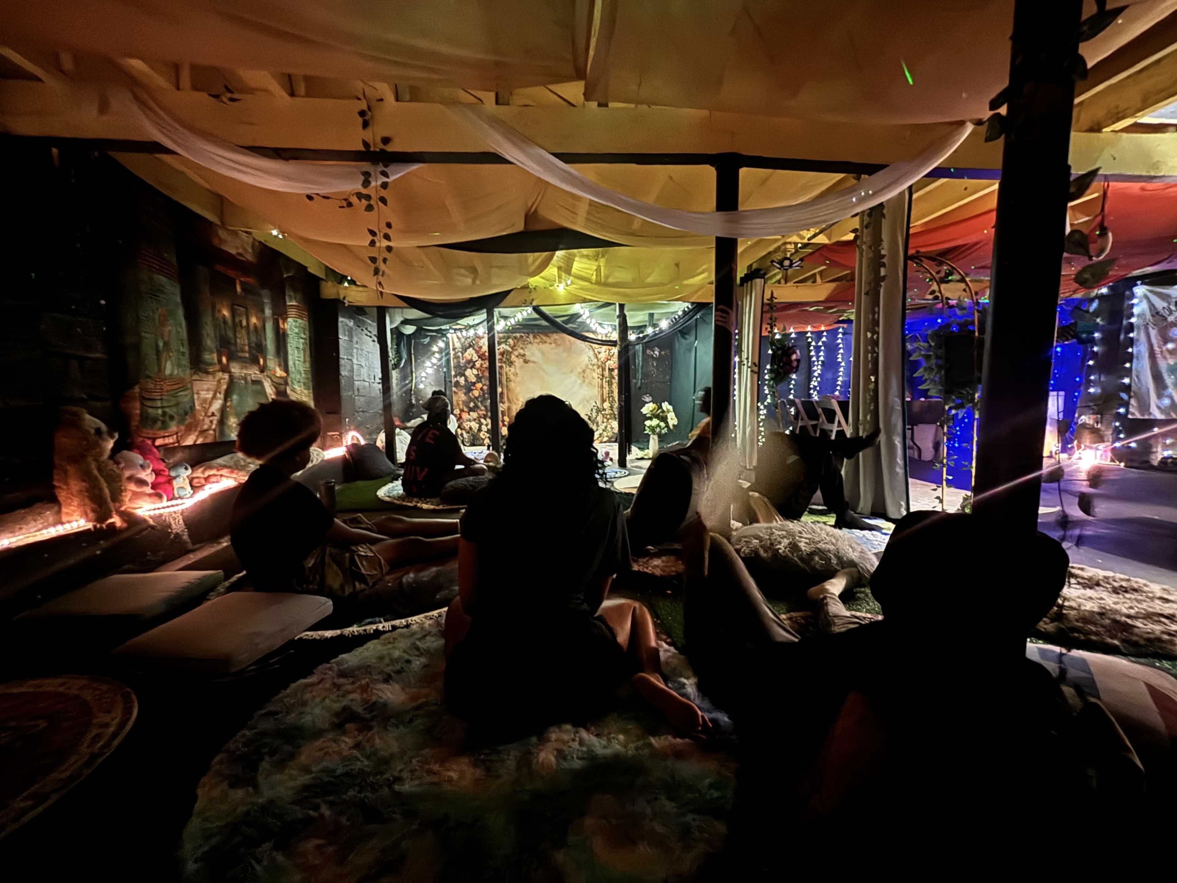 The image shows a dimly lit space with several individuals seated on floor cushions surrounded by colorful fabric drapes and ambient lighting.