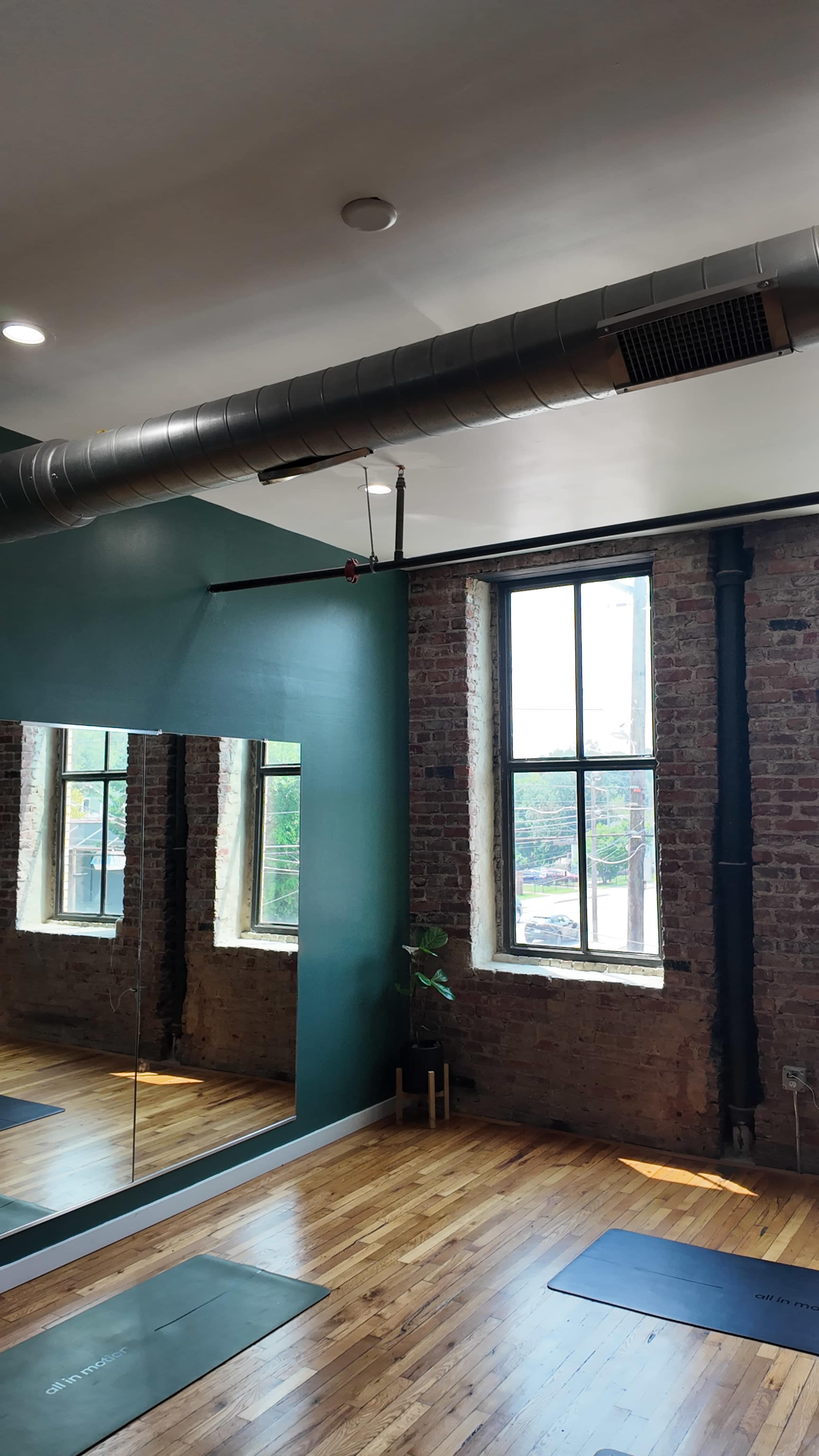 The image shows a spacious room with exposed brick walls, large windows, a mirrored wall, and wooden flooring.