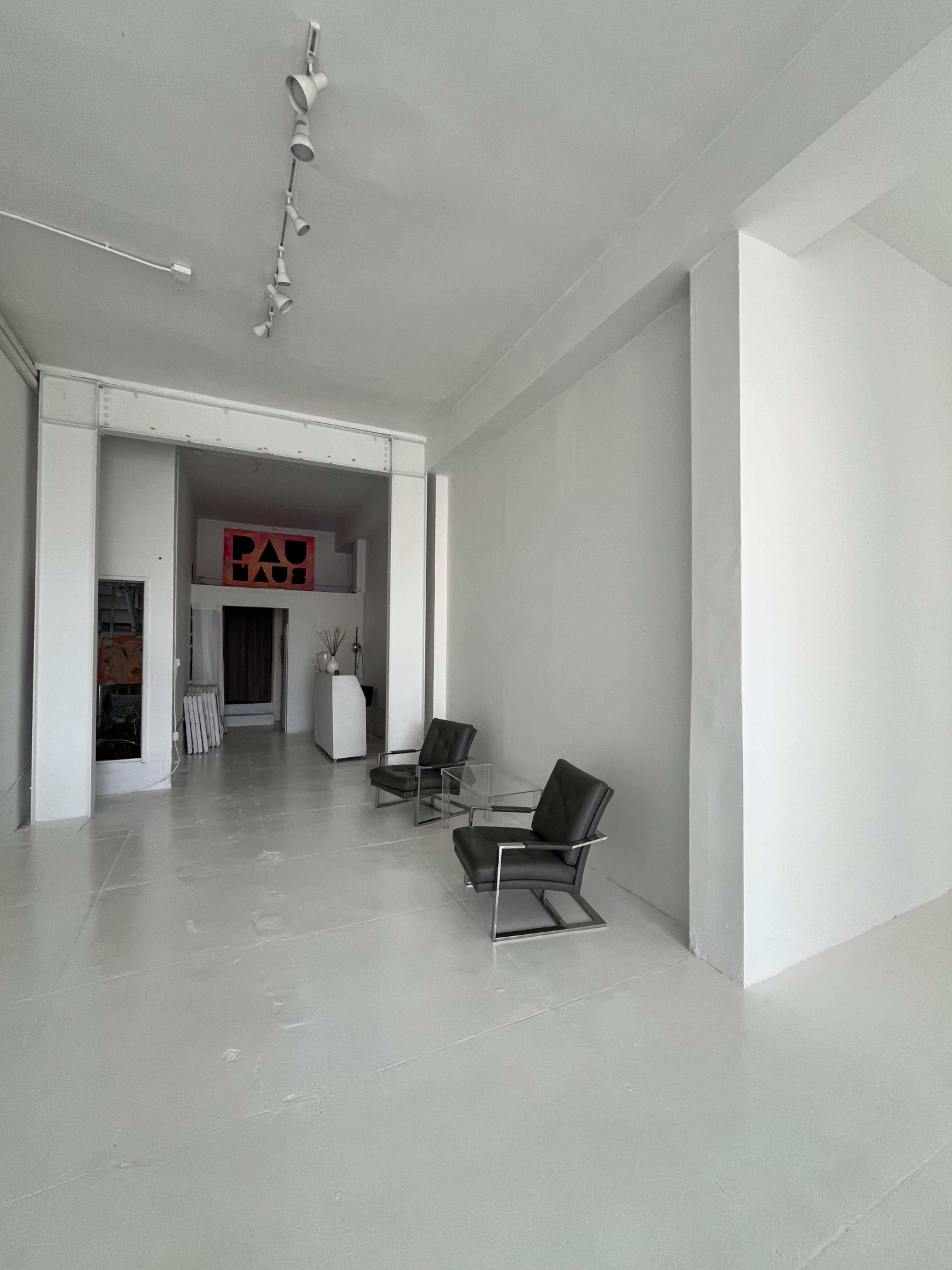 The image shows a minimalist gallery space featuring two black chairs and a glass table on a white floor, with art displayed in the background.