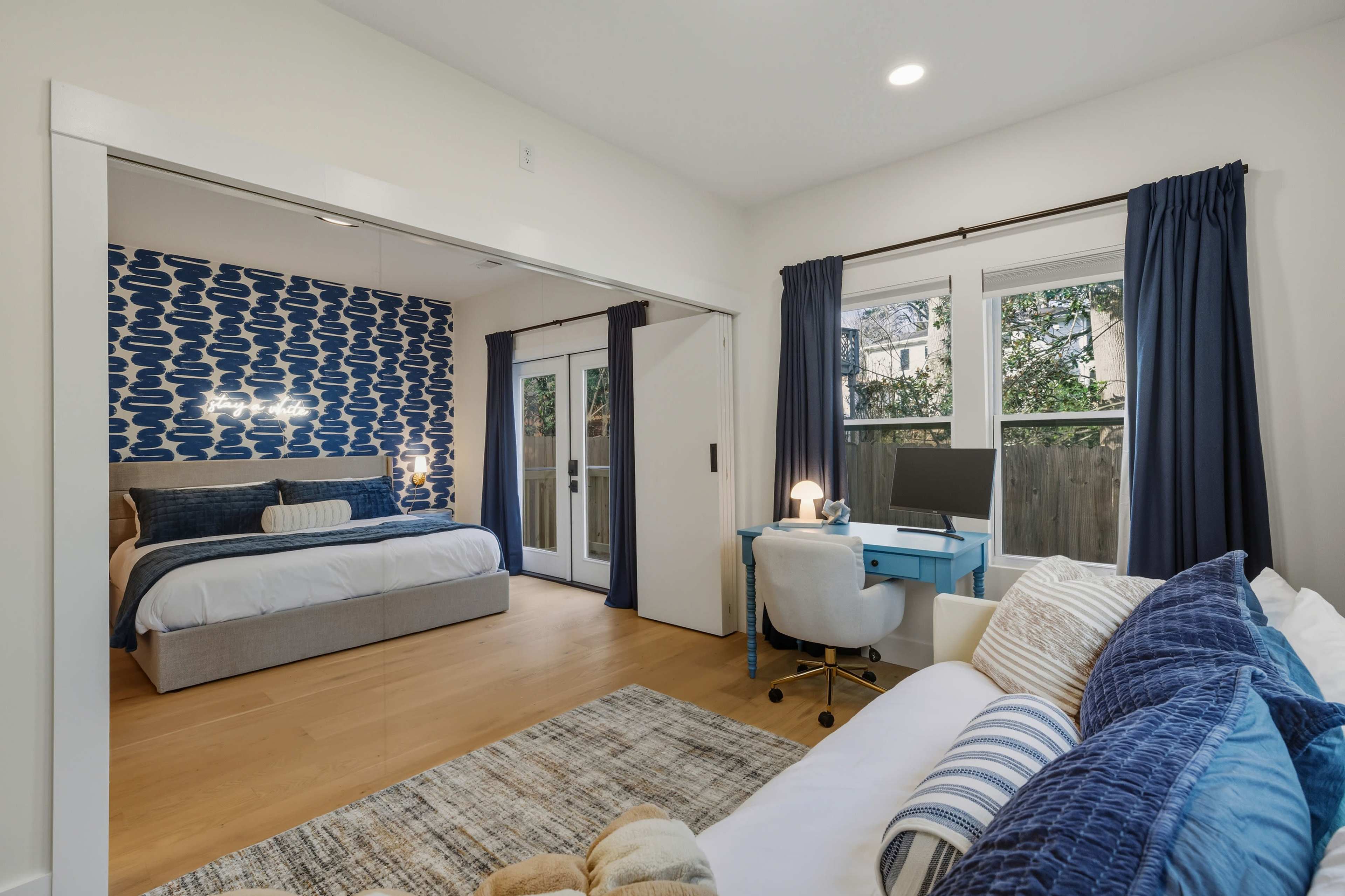 The image shows a modern bedroom with a blue and white patterned accent wall, a bed with light bedding, and a workspace featuring a desk and chair near a window.