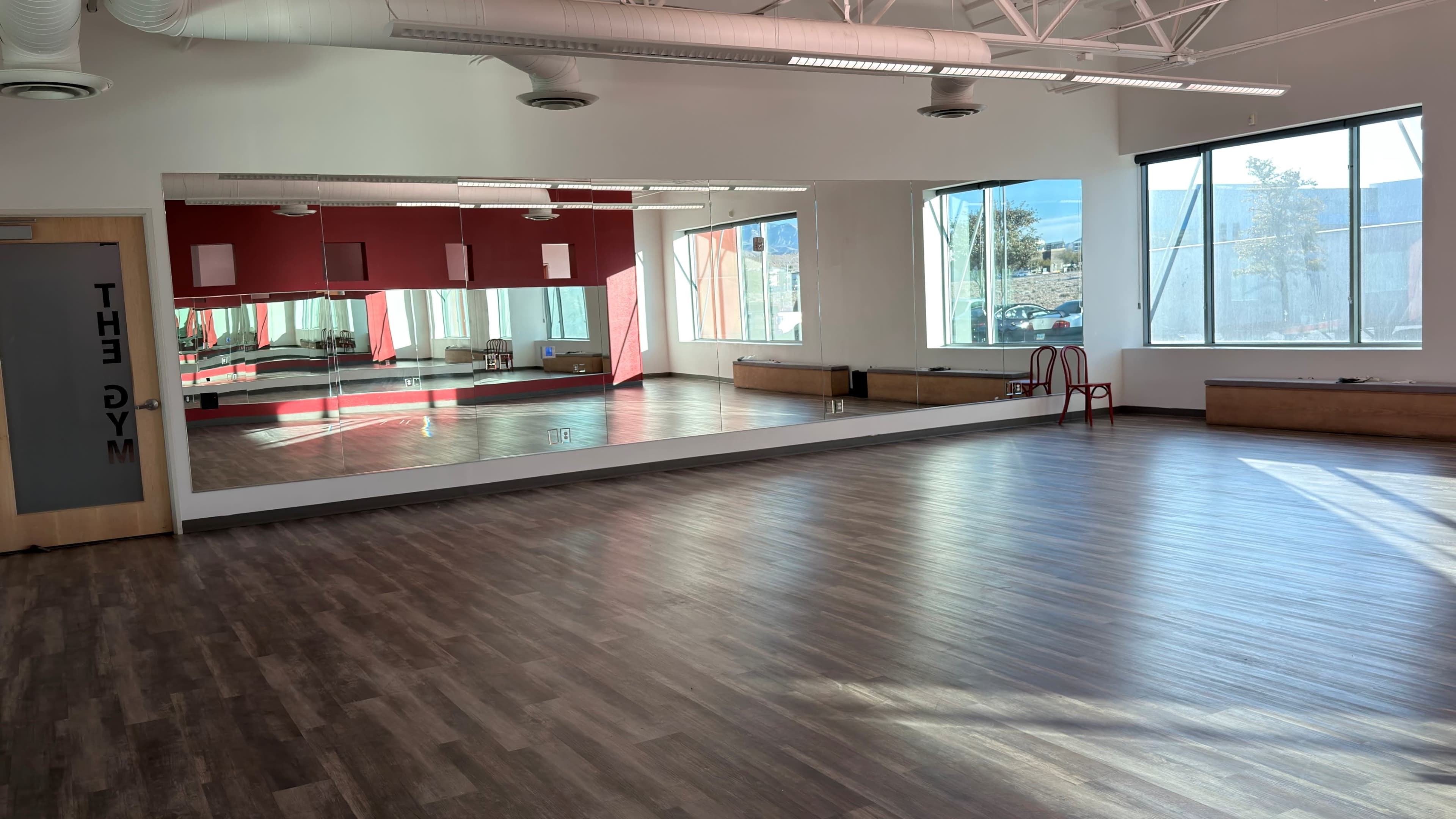 The image shows an empty gym studio with wooden floors, large windows, and a mirrored wall.