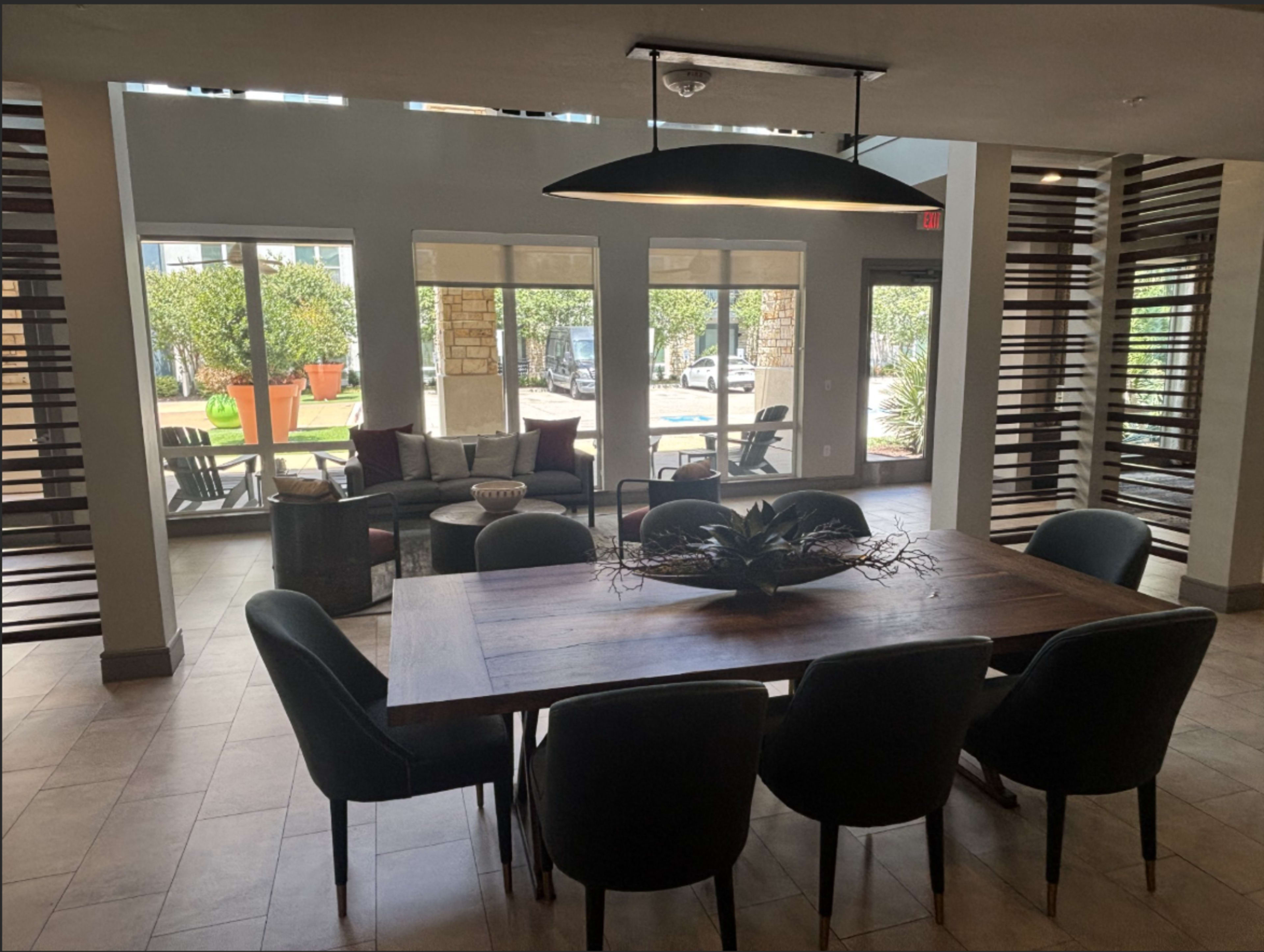 The image shows a modern communal dining area with a large wooden table surrounded by dark green chairs, featuring large windows that let in natural light and a view of an outdoor seating area.