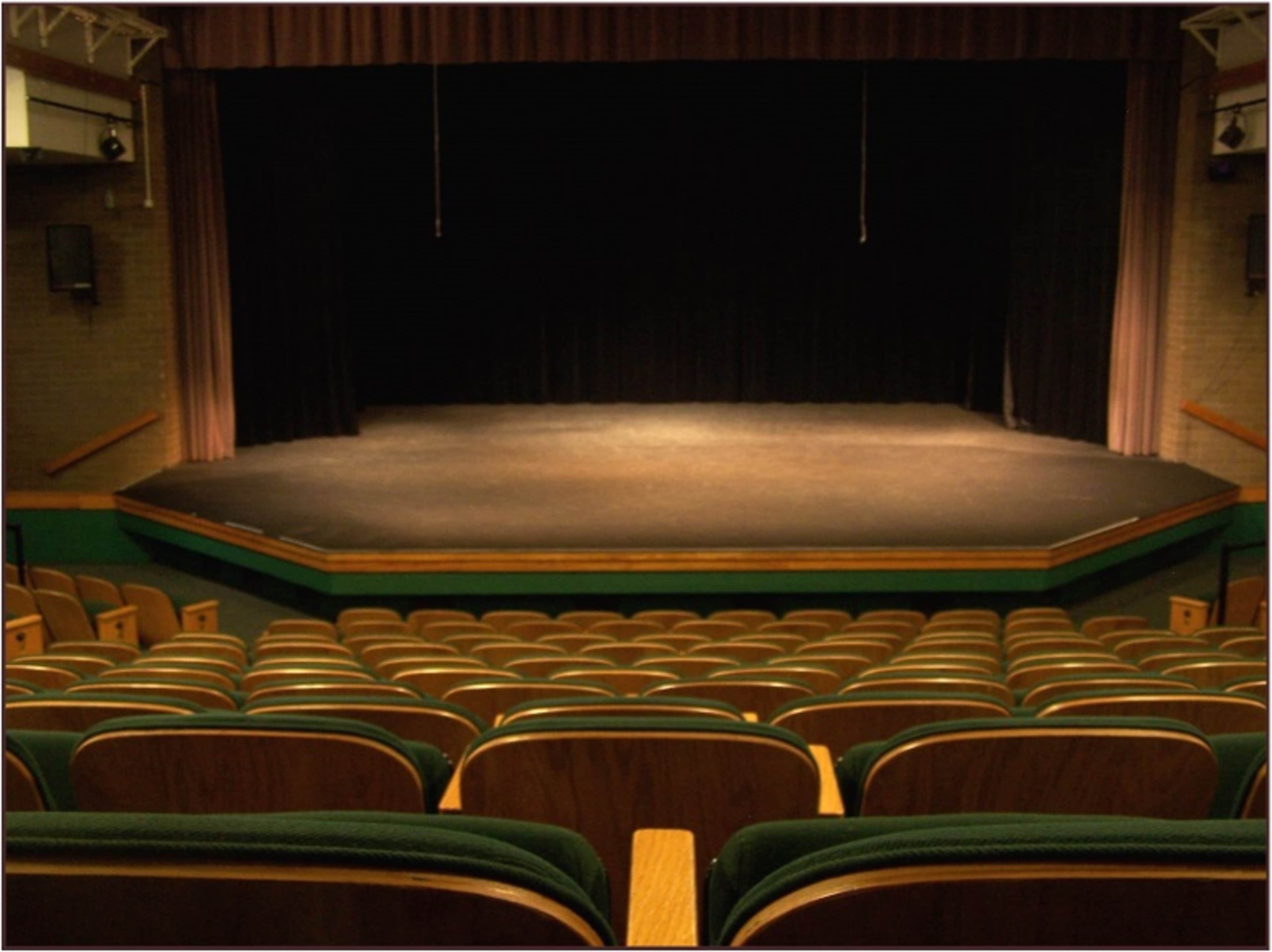 A darkened theater stage with a simple black curtain and rows of empty wooden seats in the foreground.