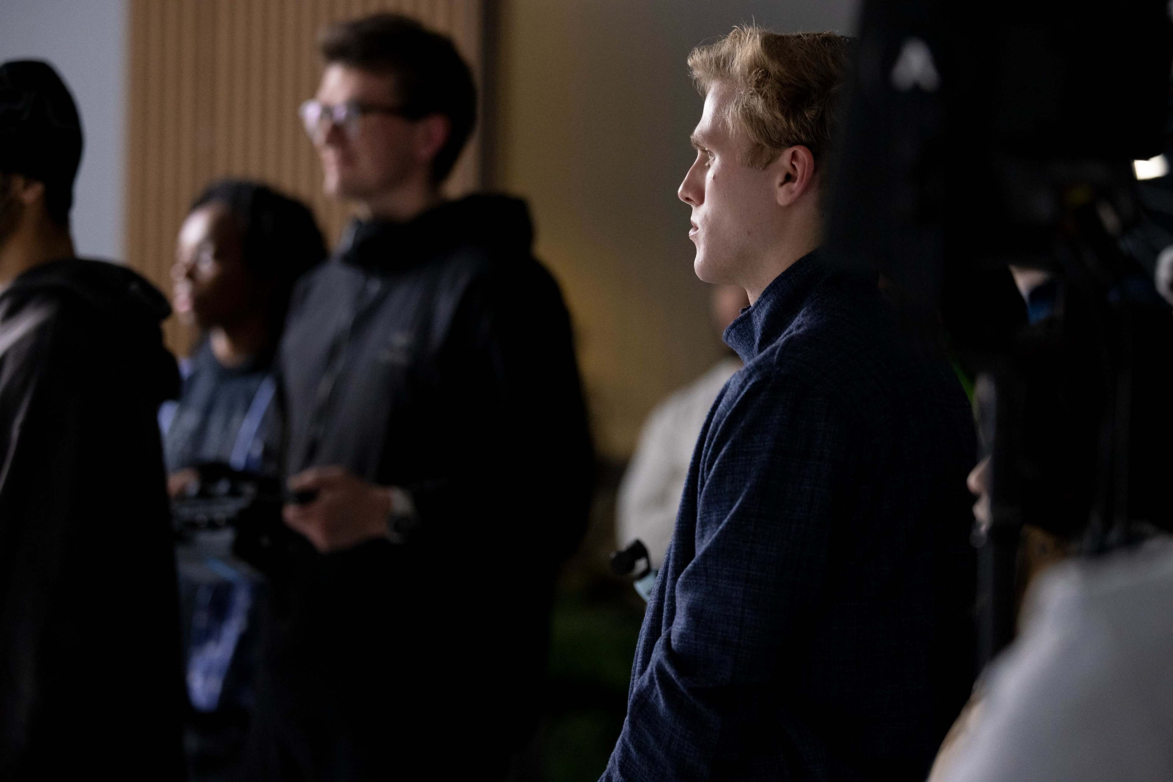 A group of people stands in a dimly lit setting, with one person in the foreground looking intently to the side.