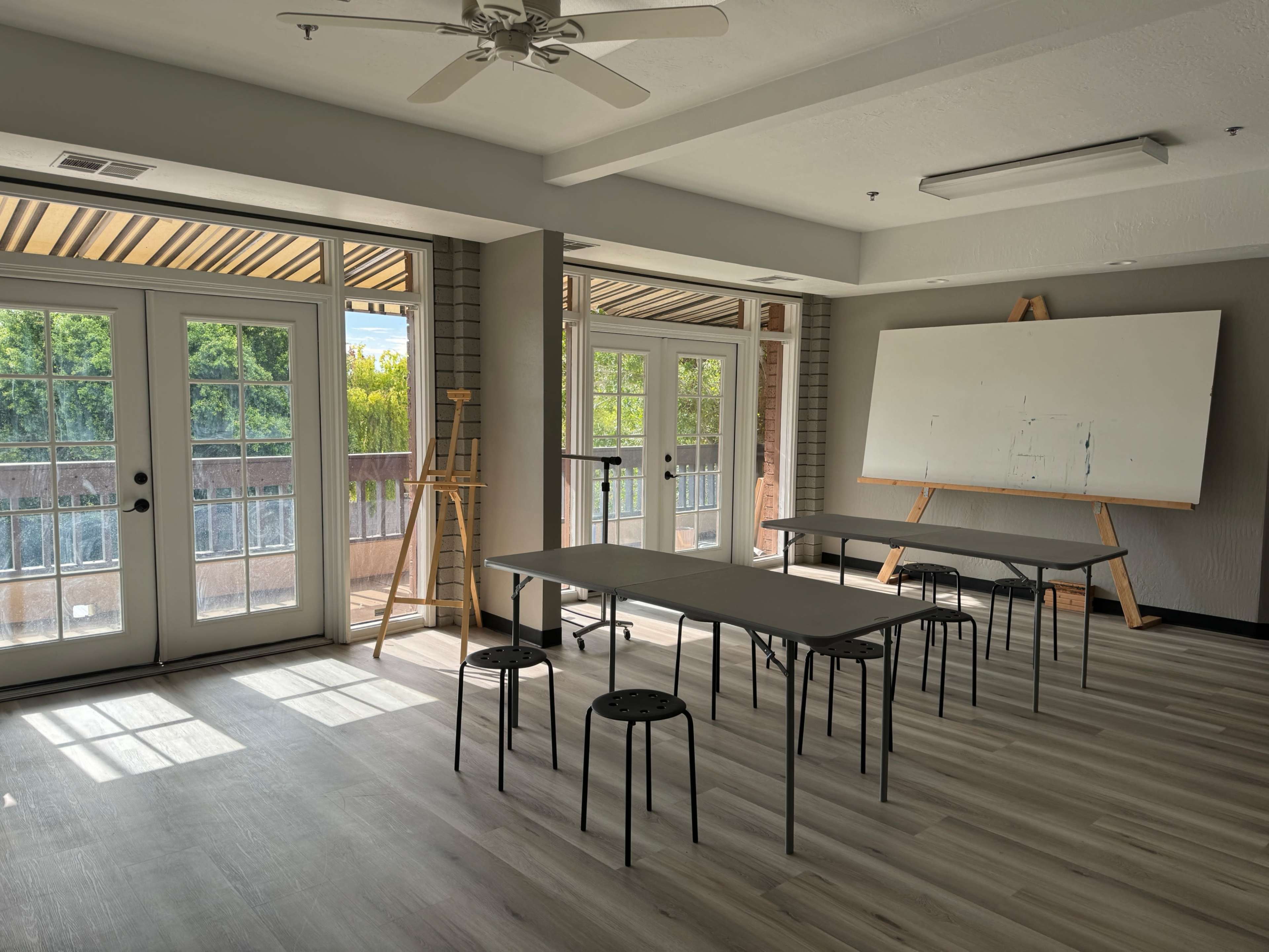 The image shows a bright, empty classroom with several tables and chairs arranged, large windows allowing natural light, and an easel with a blank canvas against one wall.