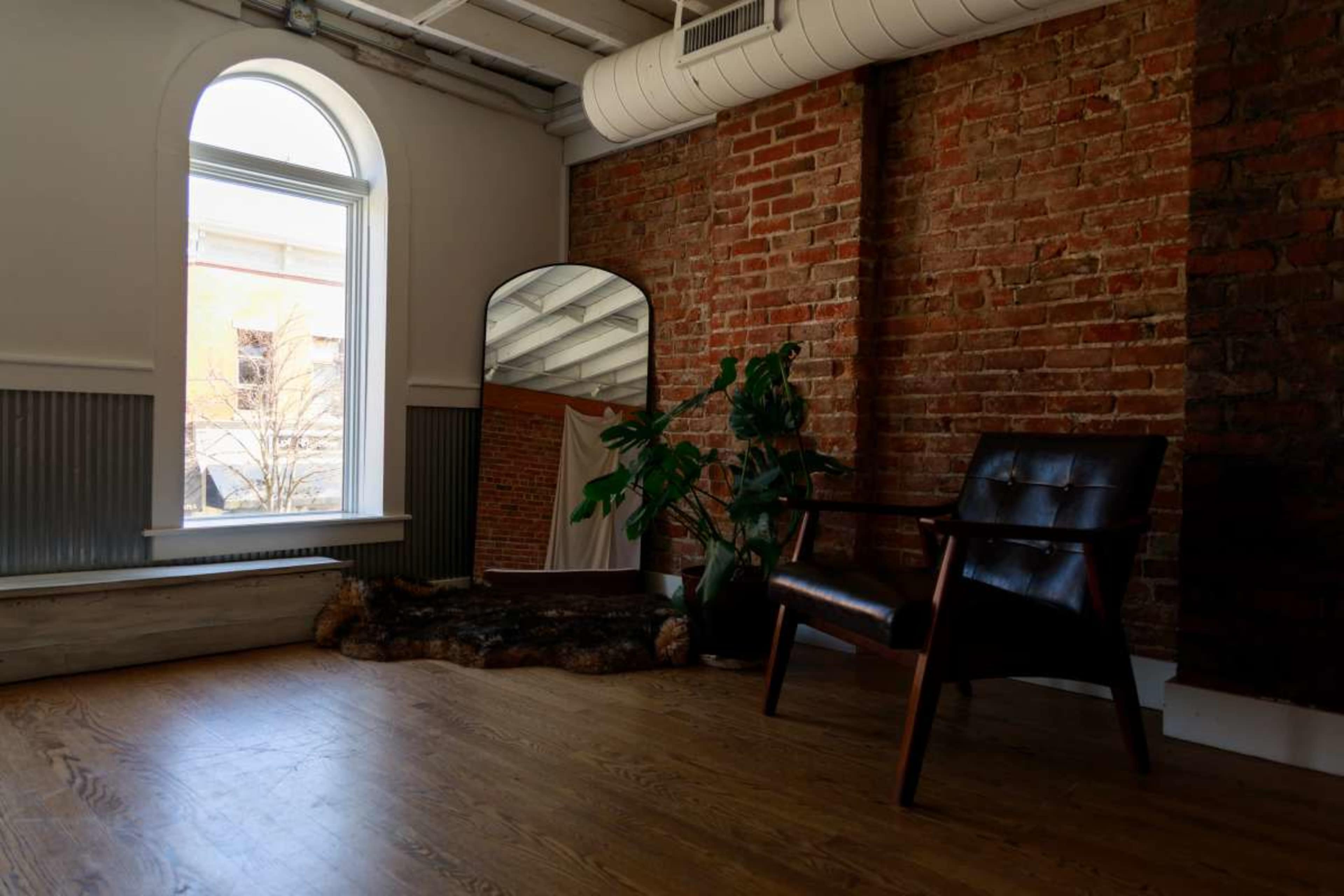 The image shows a spacious interior with a brick wall, a large mirror, a wooden chair, and a potted plant near a window.