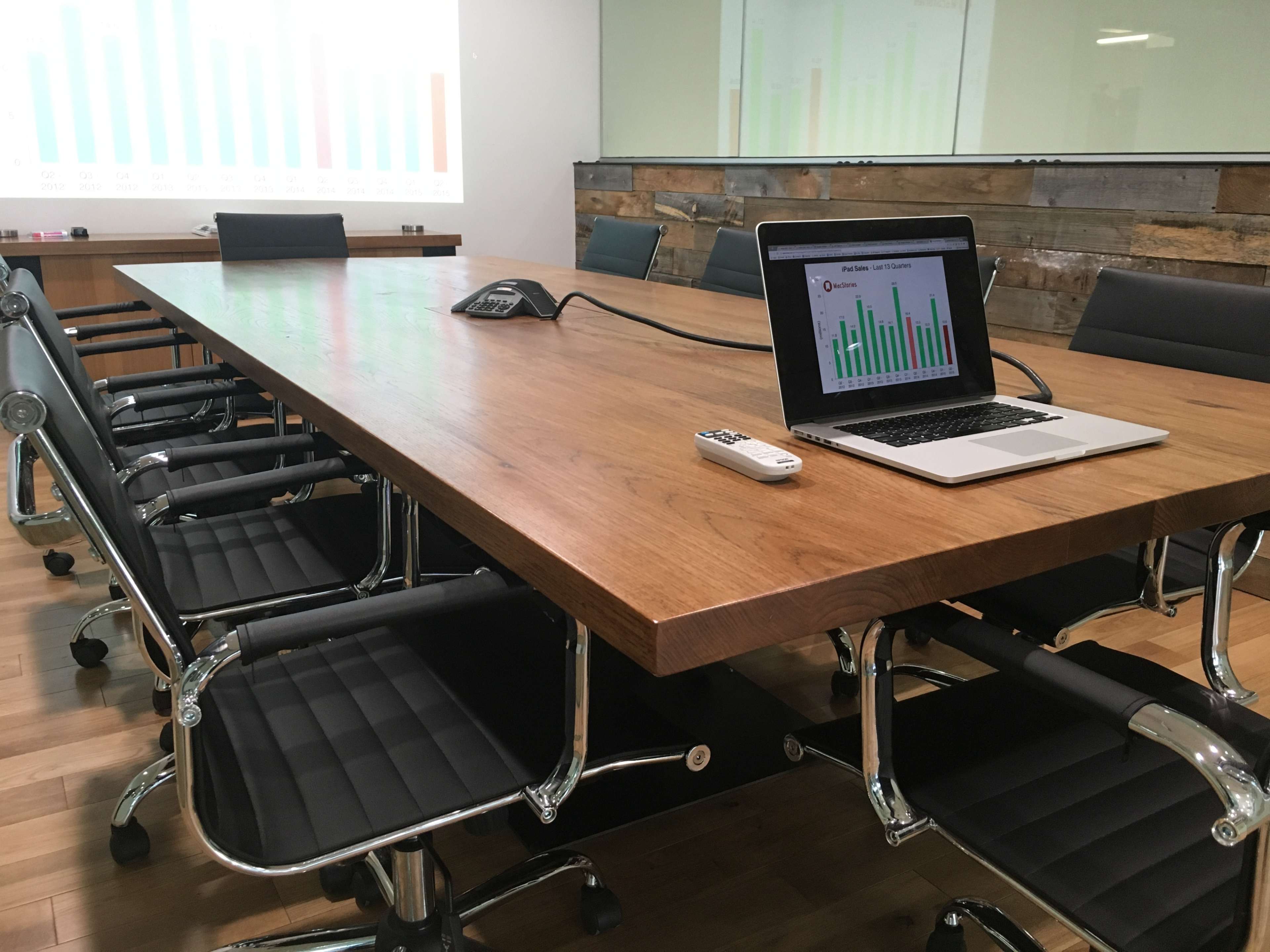 A conference room table is set with a laptop displaying a bar graph and several black rolling chairs arranged around it, with a projector screen in the background.