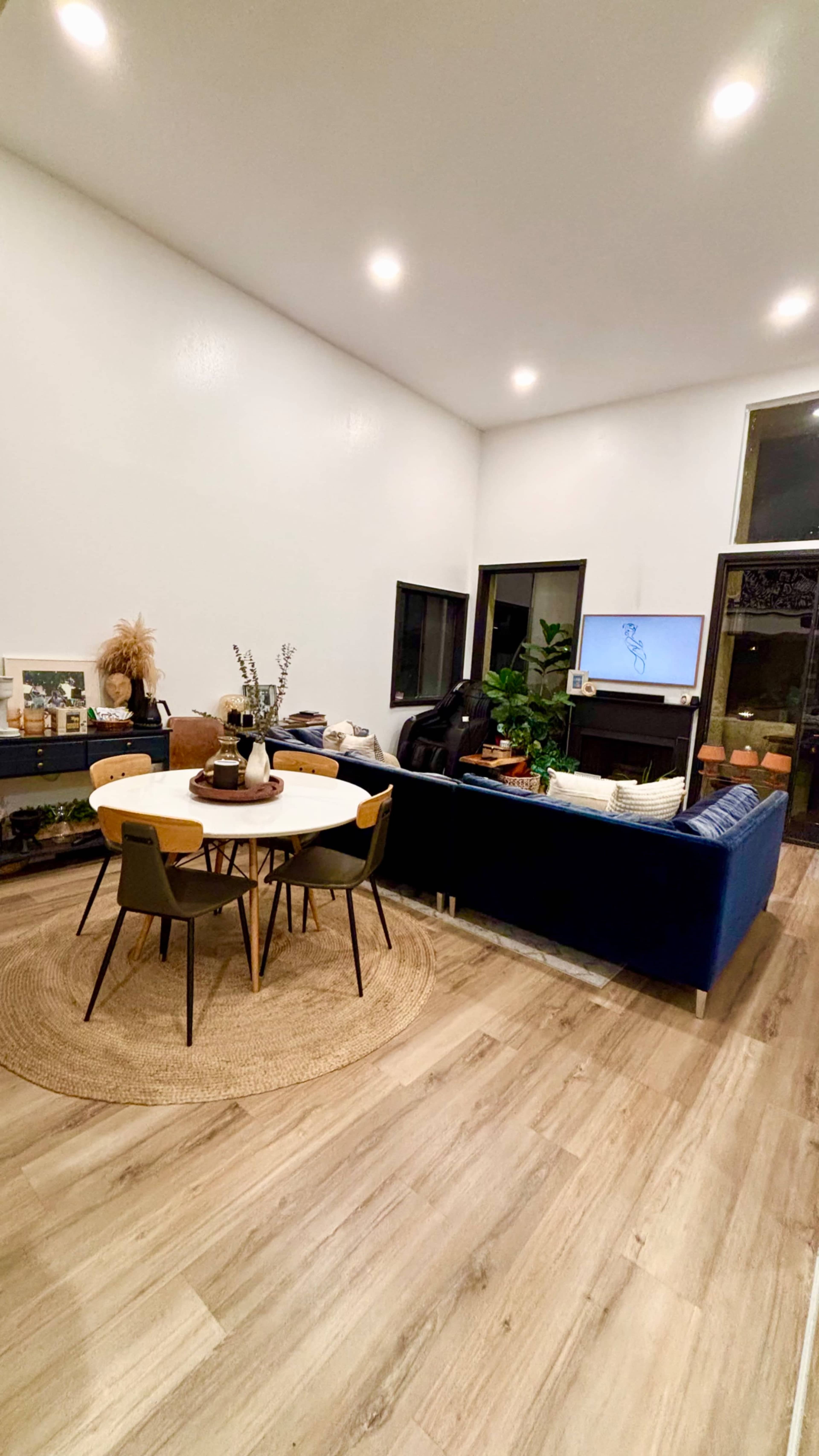 The image shows a modern living room with a circular dining table, black chairs, a blue sofa, and large windows.