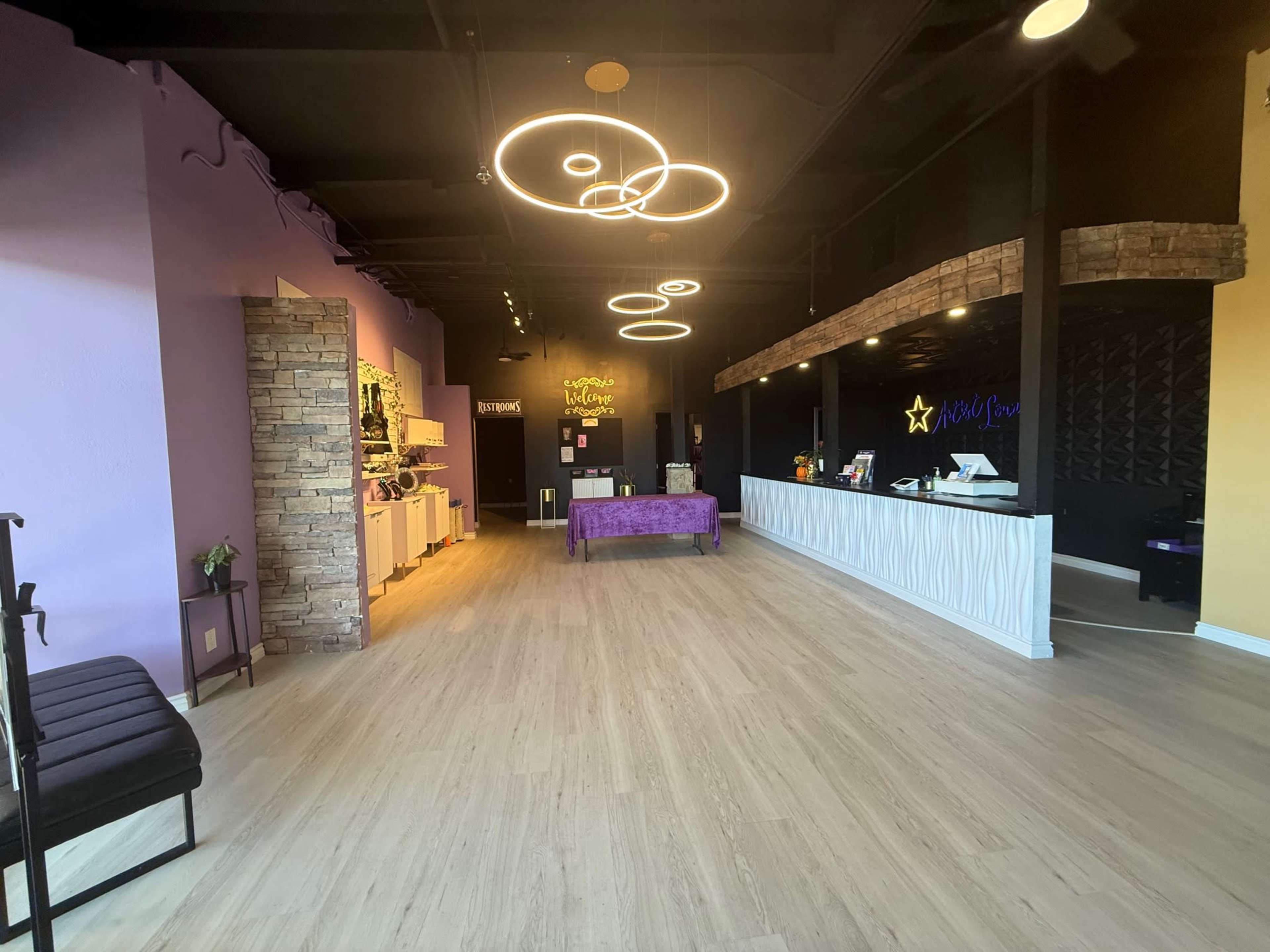 The image shows an interior of a brightly lit salon or spa with a reception desk, a purple accent wall, and modern lighting fixtures.