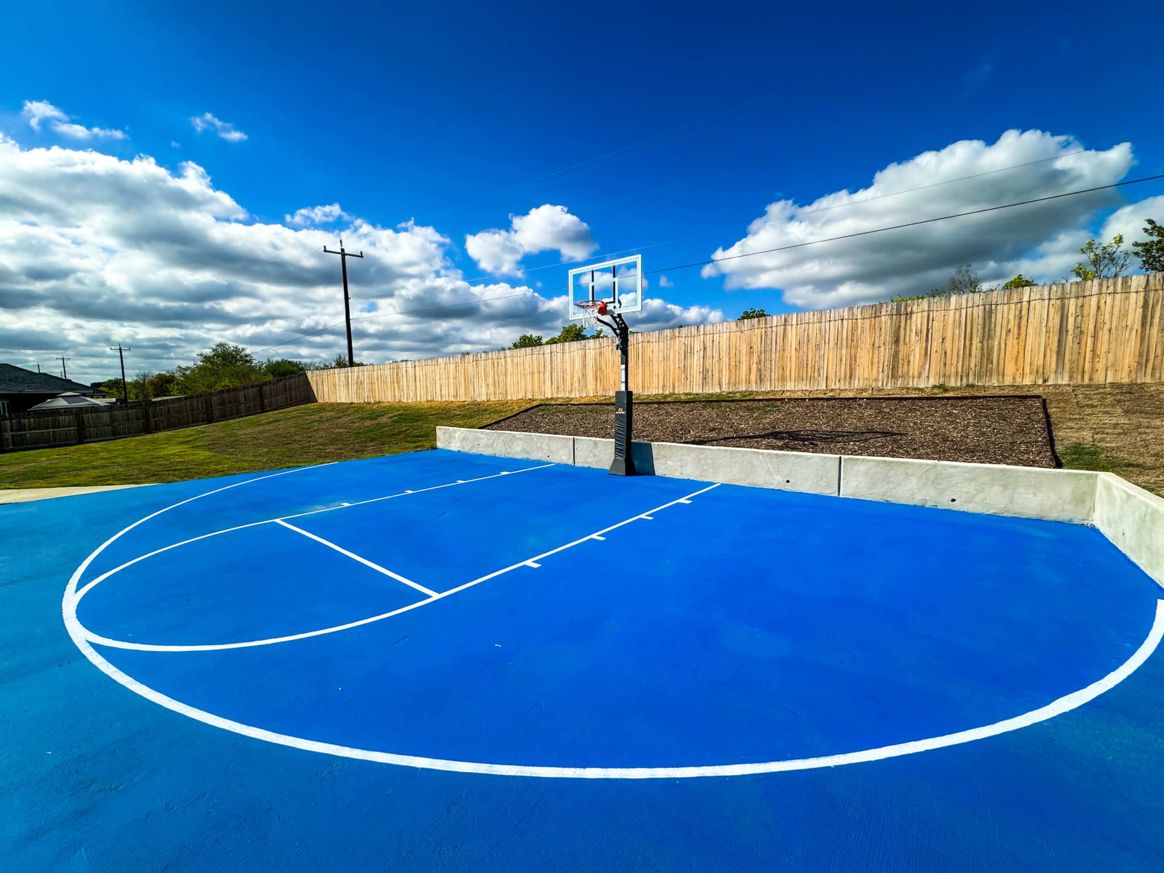 The image shows a blue outdoor basketball court with a hoop and a grassy area enclosed by a wooden fence.
