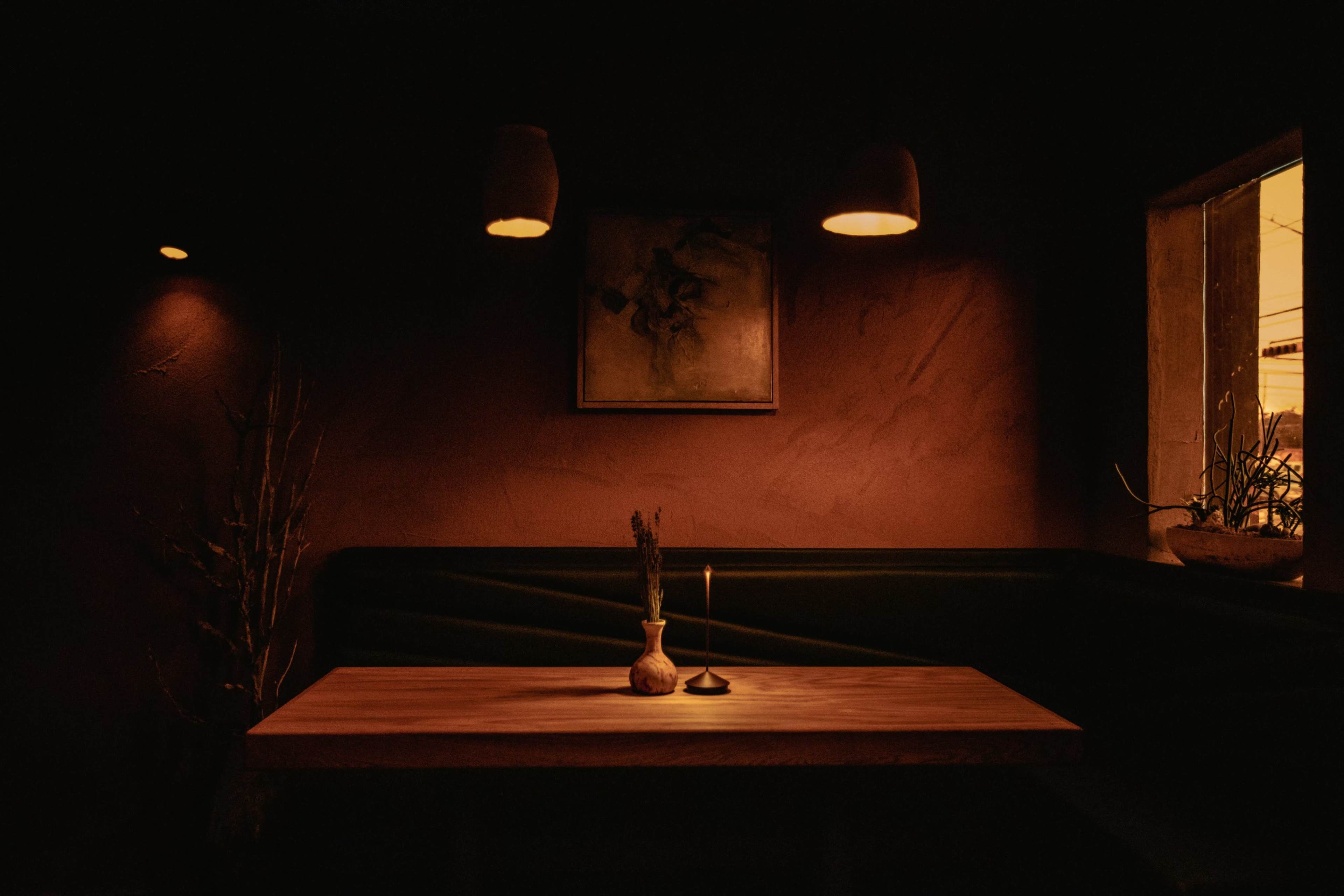 A wooden table with a vase and candle is positioned in a dimly lit corner of a restaurant, illuminated by warm light from hanging fixtures and a window.