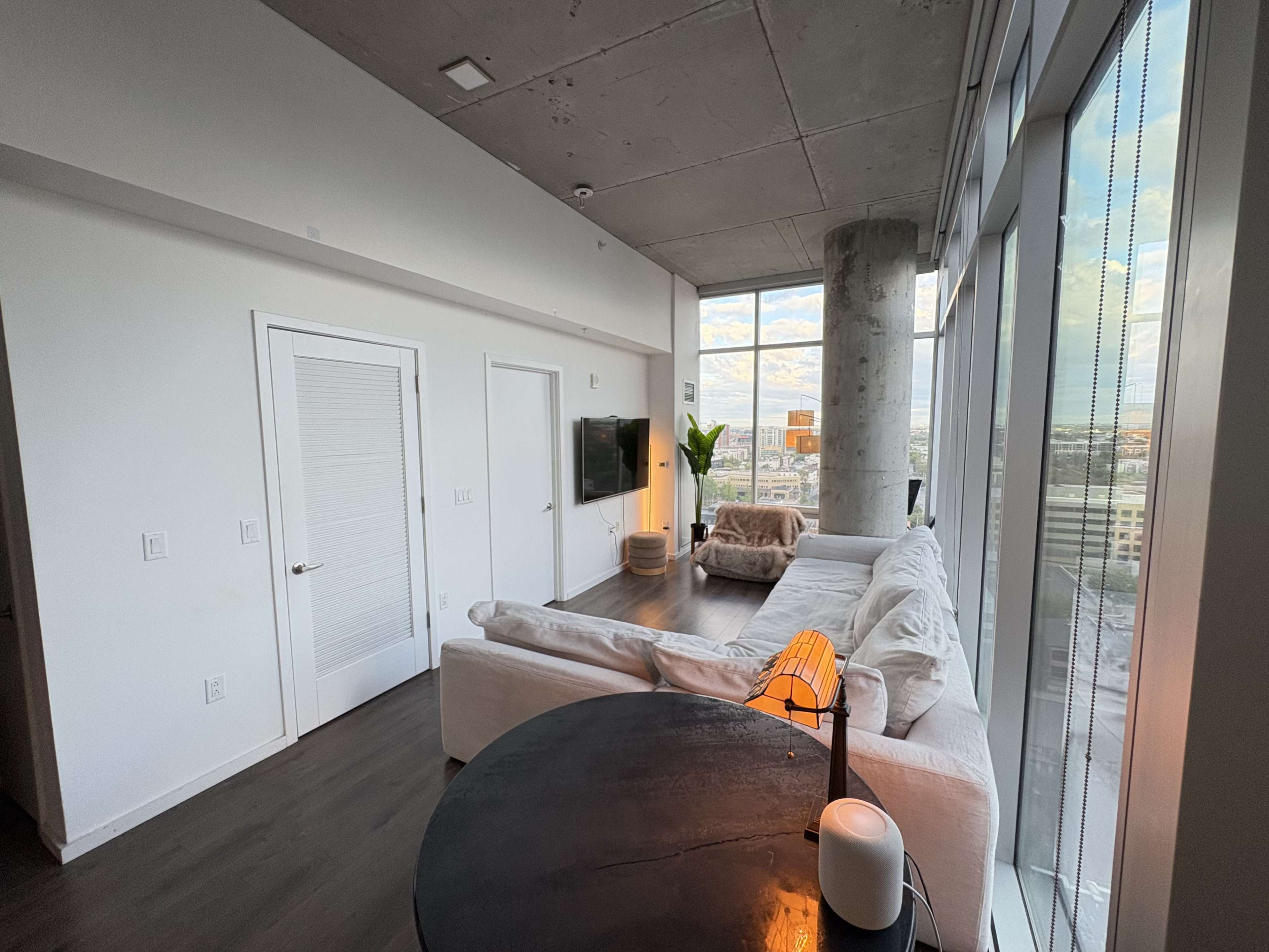 The image shows a modern apartment living area with large windows, a white sectional sofa, a round black table, and a view of the city skyline.