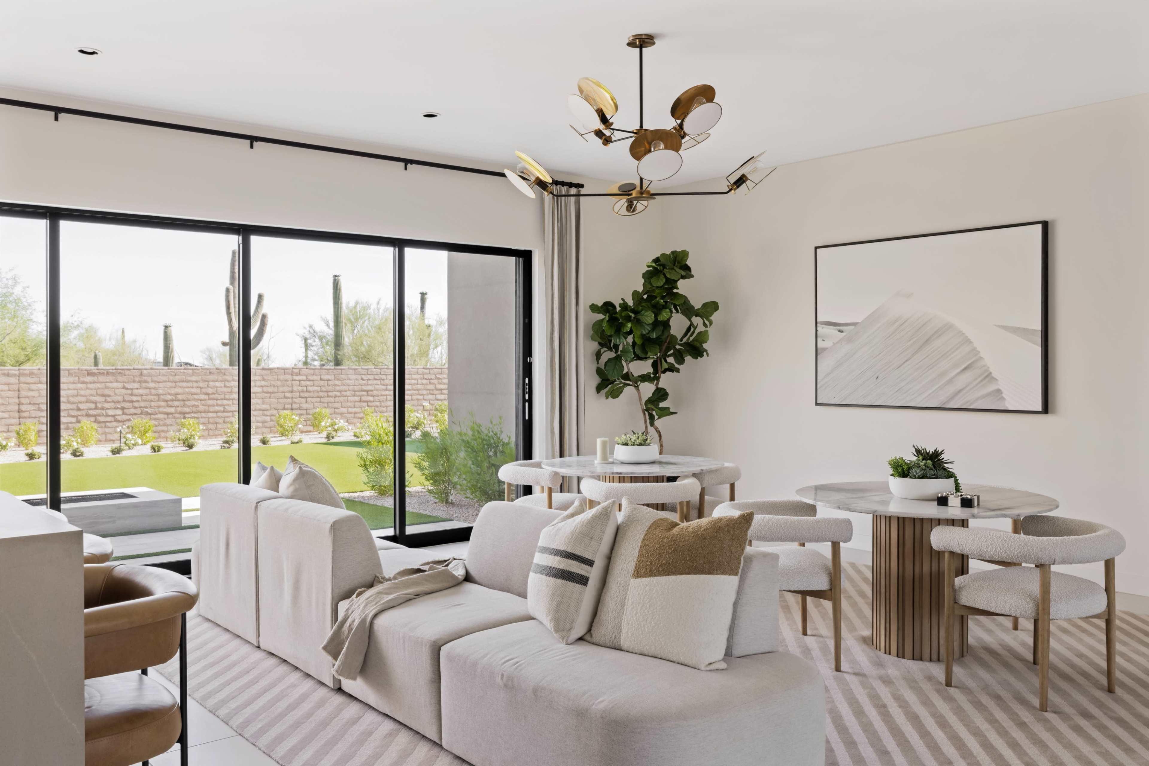 The image shows a modern living room featuring a light-colored sofa, a circular dining table, and a large window that opens to a landscaped outdoor area.