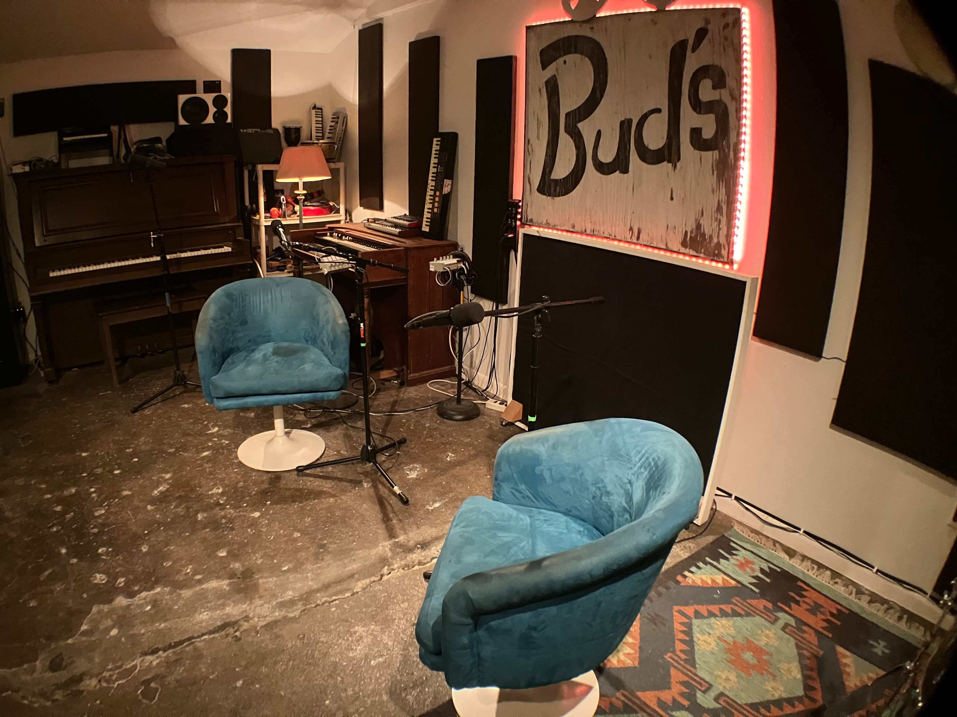 The image shows a recording studio with two blue chairs, a microphone, a piano in the background, and various musical instruments and equipment.