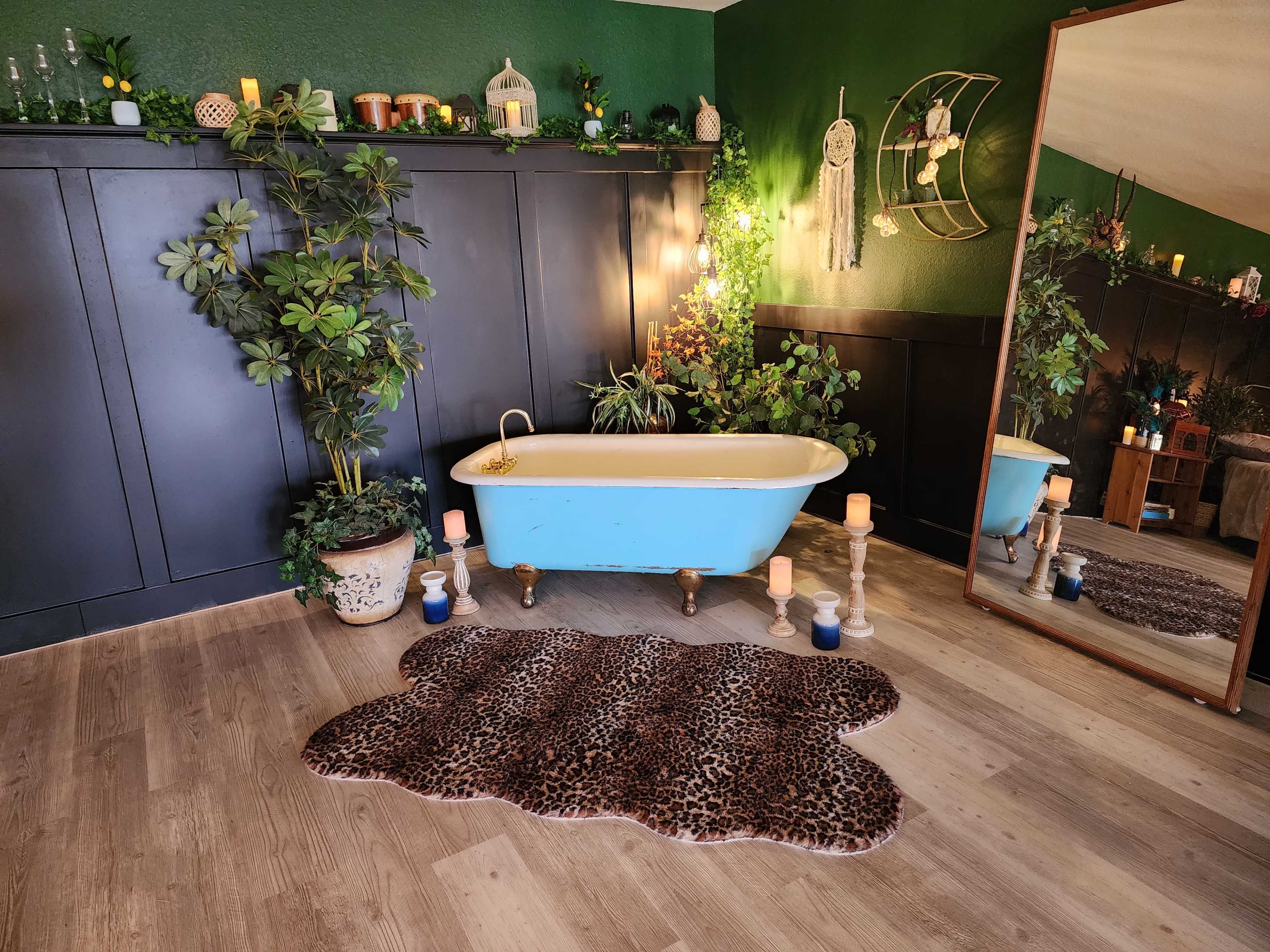 The image shows a vintage-style bathroom with a freestanding bathtub, surrounded by plants, candles, and a large mirror against a dark green wall.