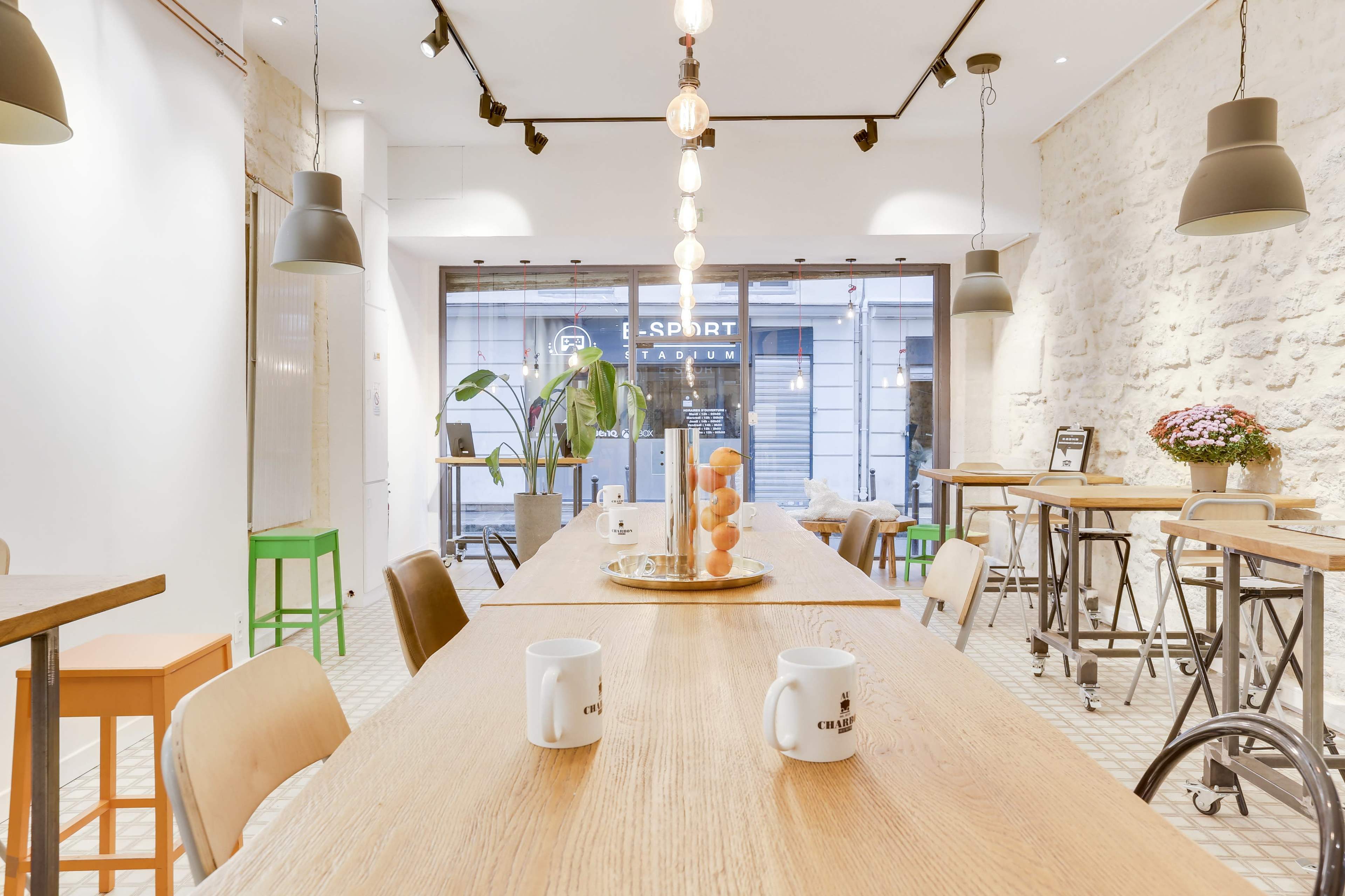 A spacious café features wooden tables, colorful bar stools, and large windows with a view of the street outside.