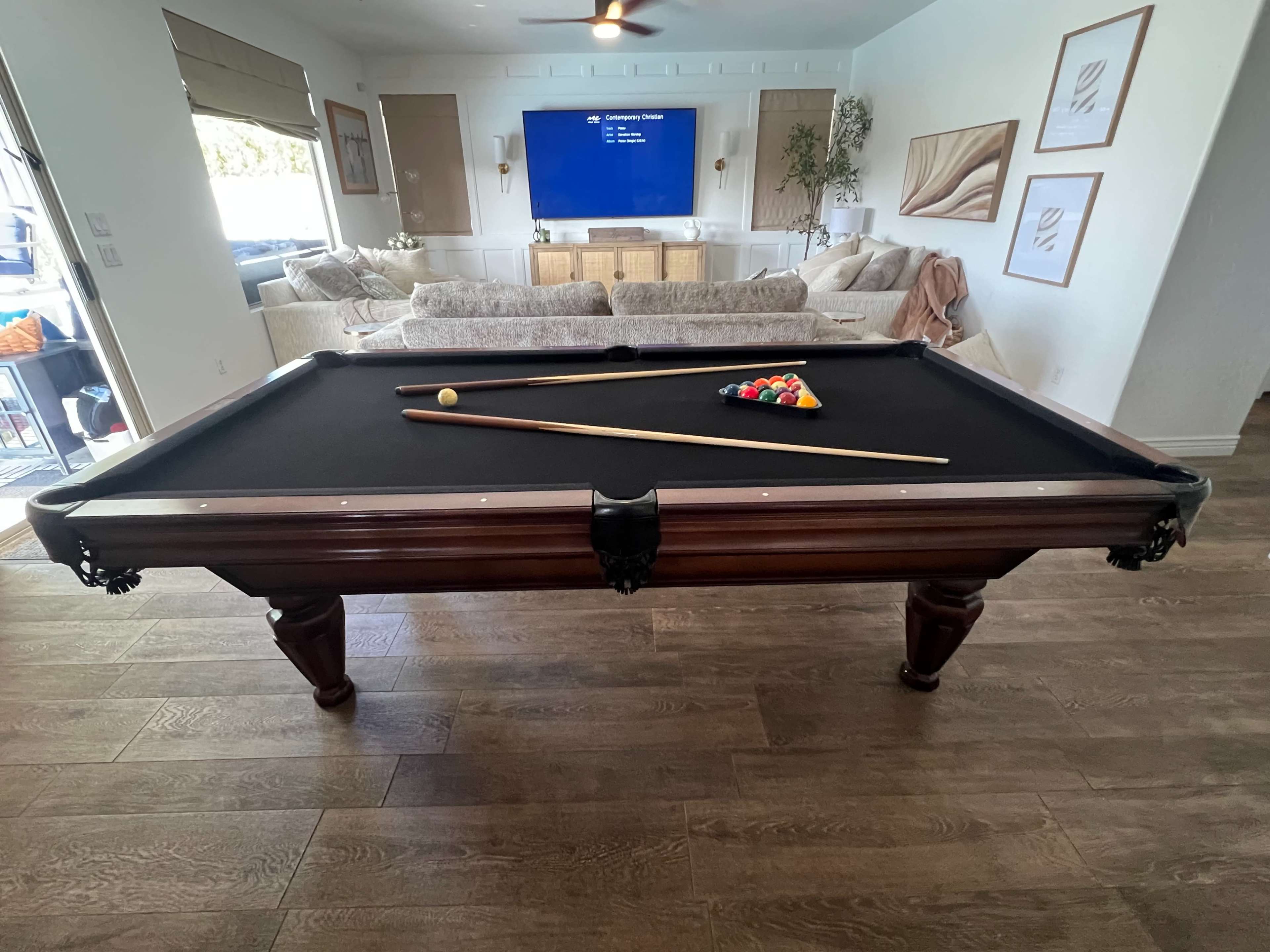A pool table with a full set of balls and cues sits in a room featuring a couch and a television in the background.