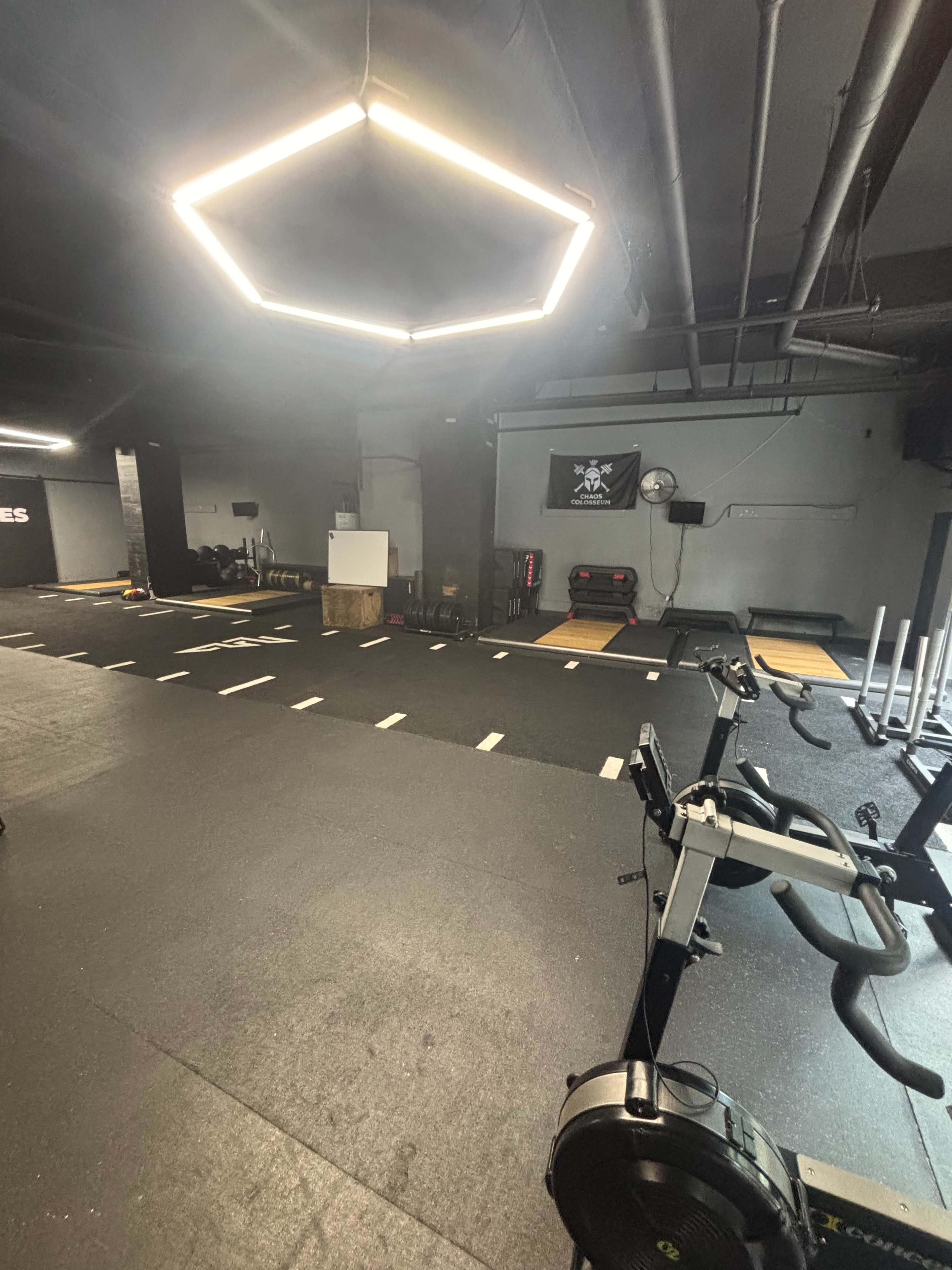 The image shows a dimly lit gym with black rubber flooring, various fitness equipment, and a modern hexagonal light fixture hanging from the ceiling.