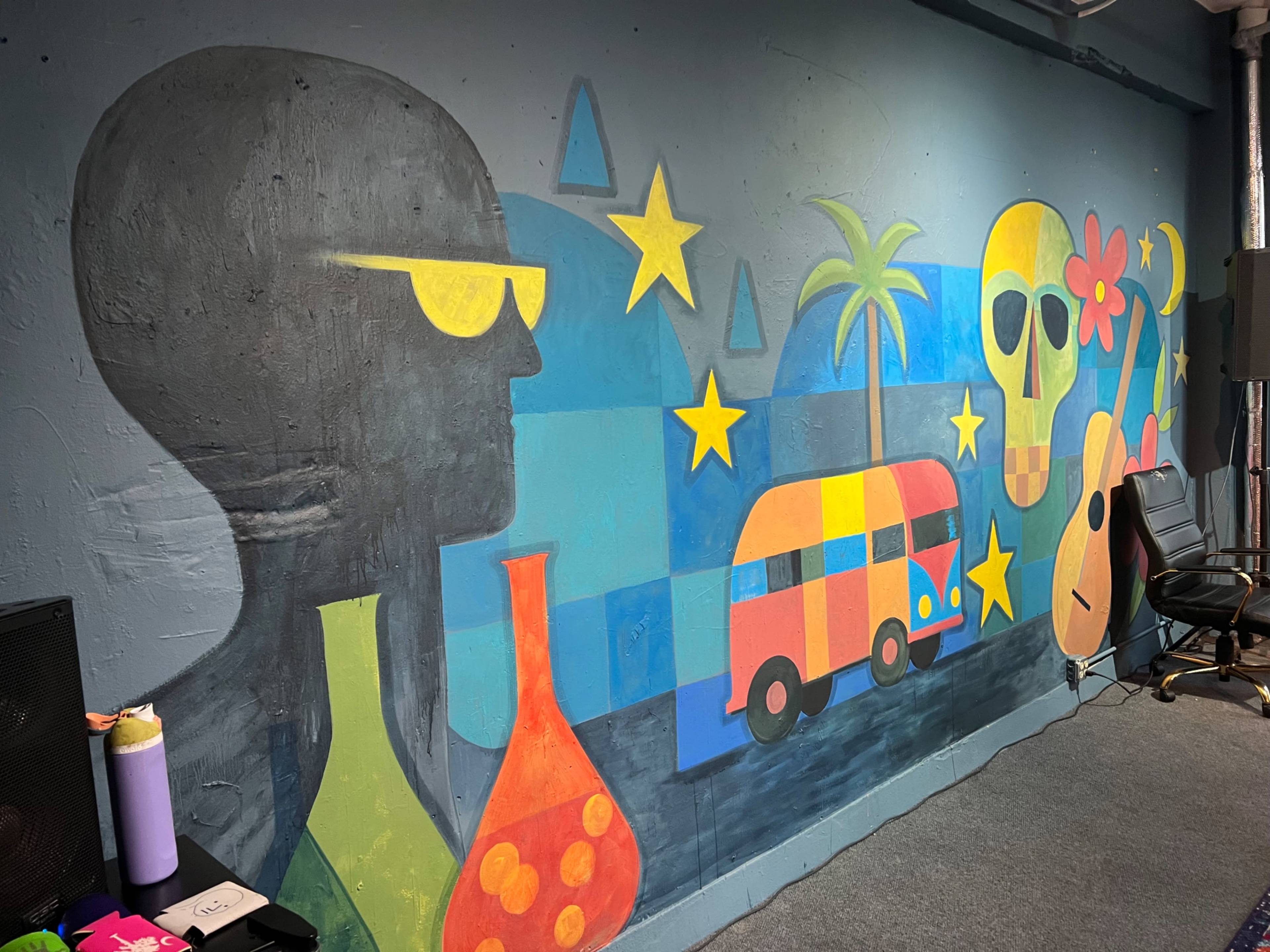 The image features a colorful mural depicting abstract shapes, a silhouette wearing sunglasses, flowers, stars, a guitar, and a van against a blue background.