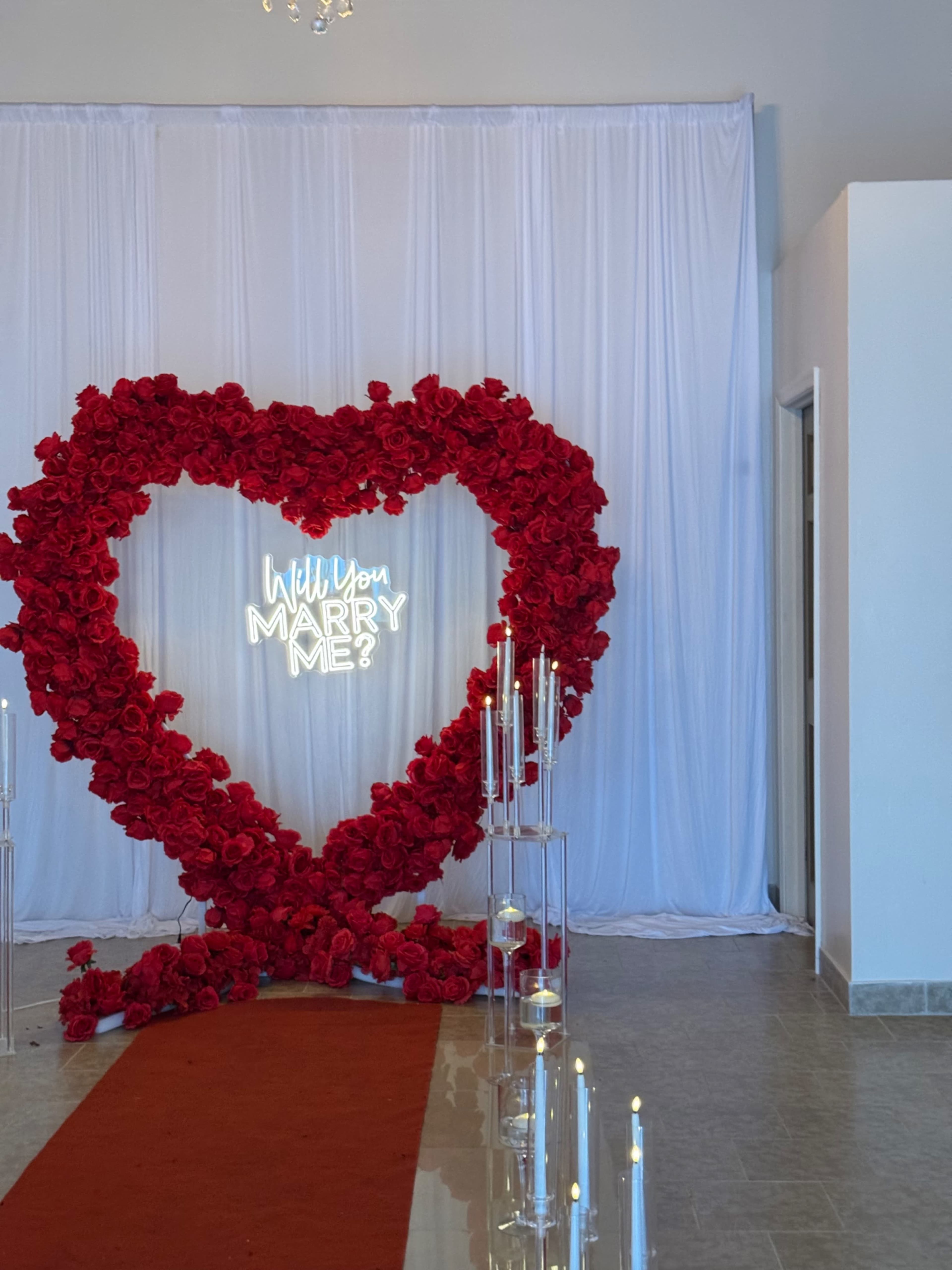 A large heart-shaped arrangement of red roses is set in front of a white backdrop, with the words "Will You Marry Me?" illuminated in neon and surrounded by candles along a red carpet.
