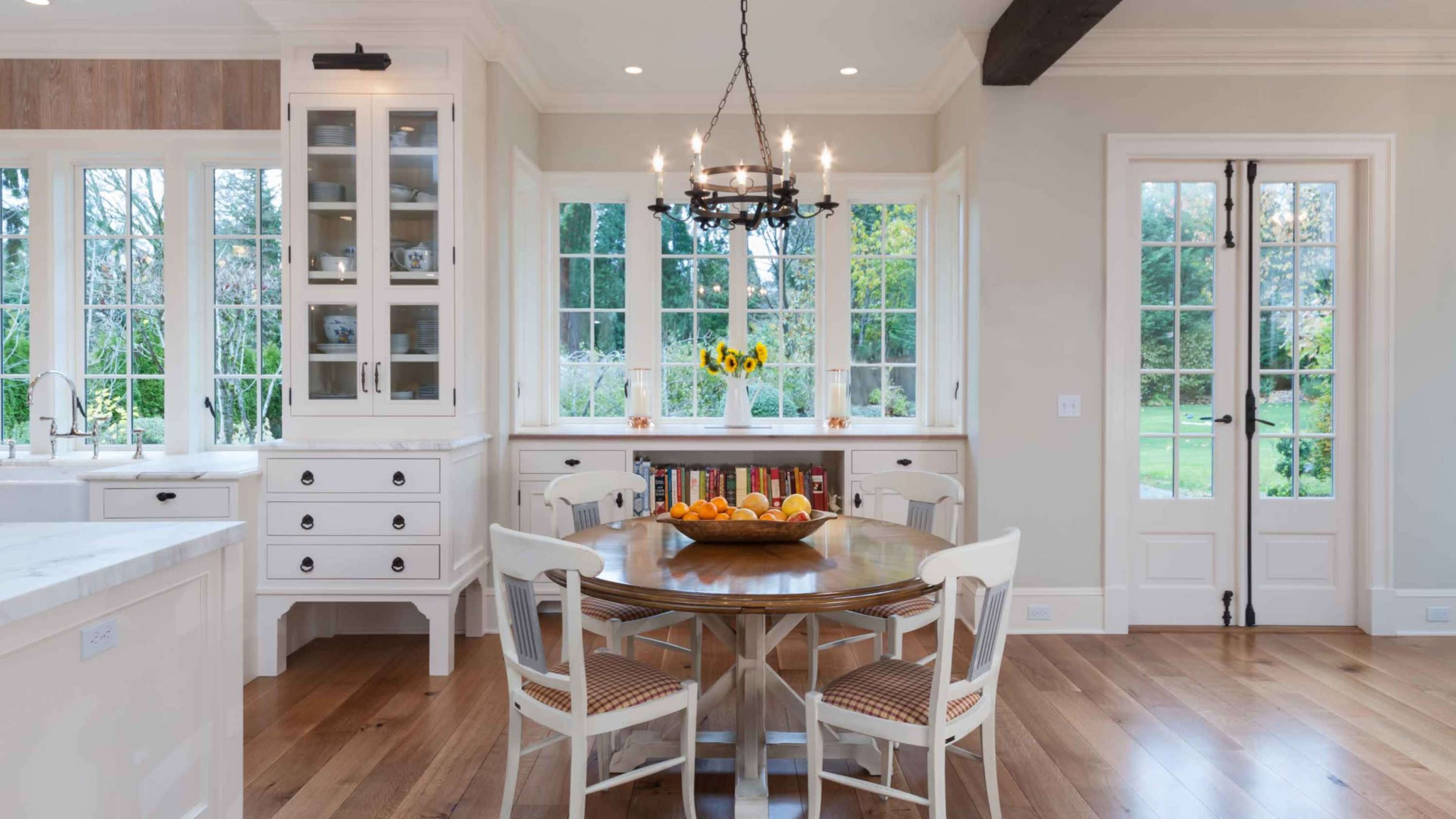 A bright kitchen features a round dining table with a fruit bowl, surrounded by white chairs, and large windows that overlook a garden.