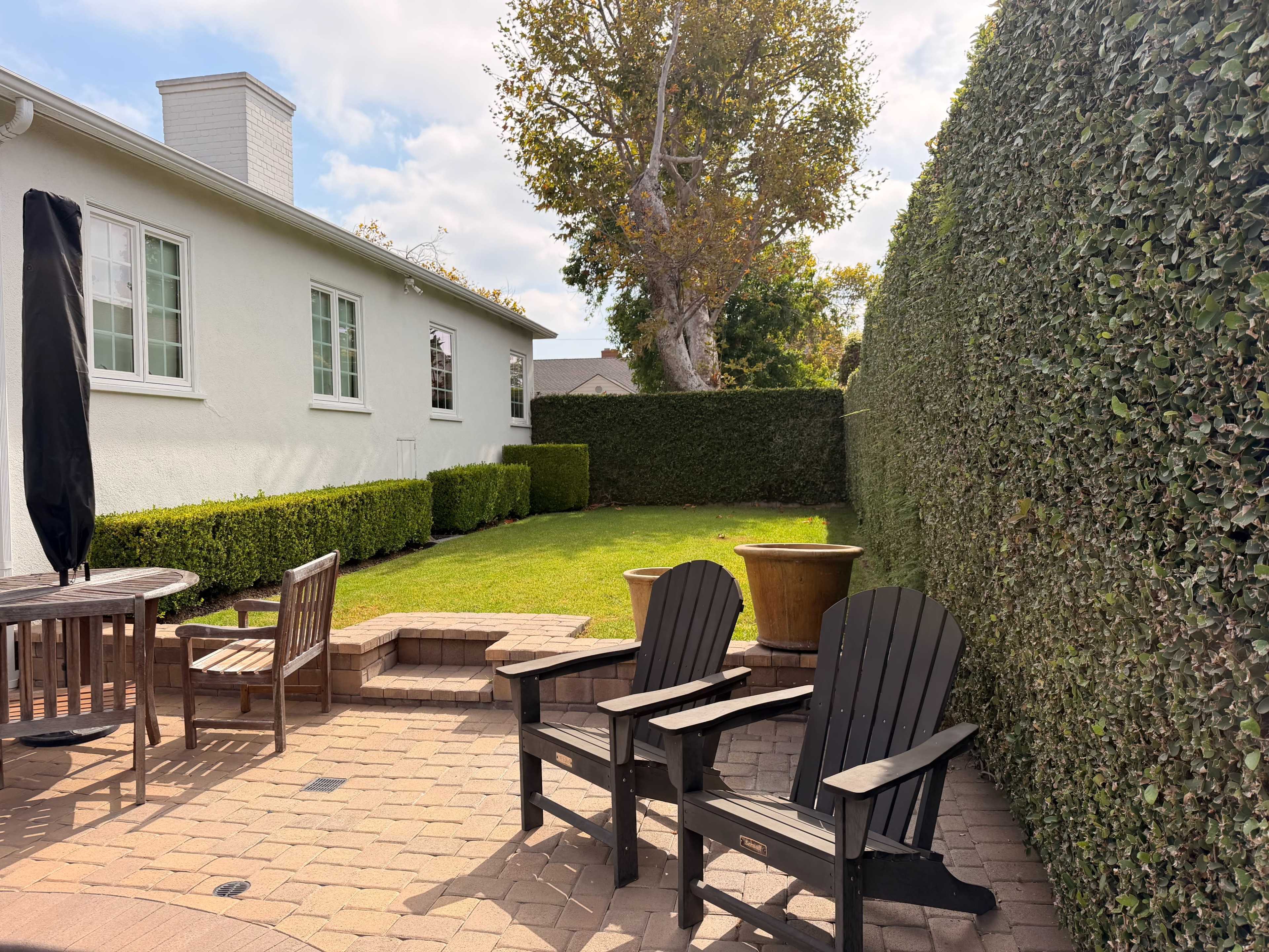 The image shows a well-manicured backyard featuring a patio with black Adirondack chairs, a circular planter, and neatly trimmed hedges.