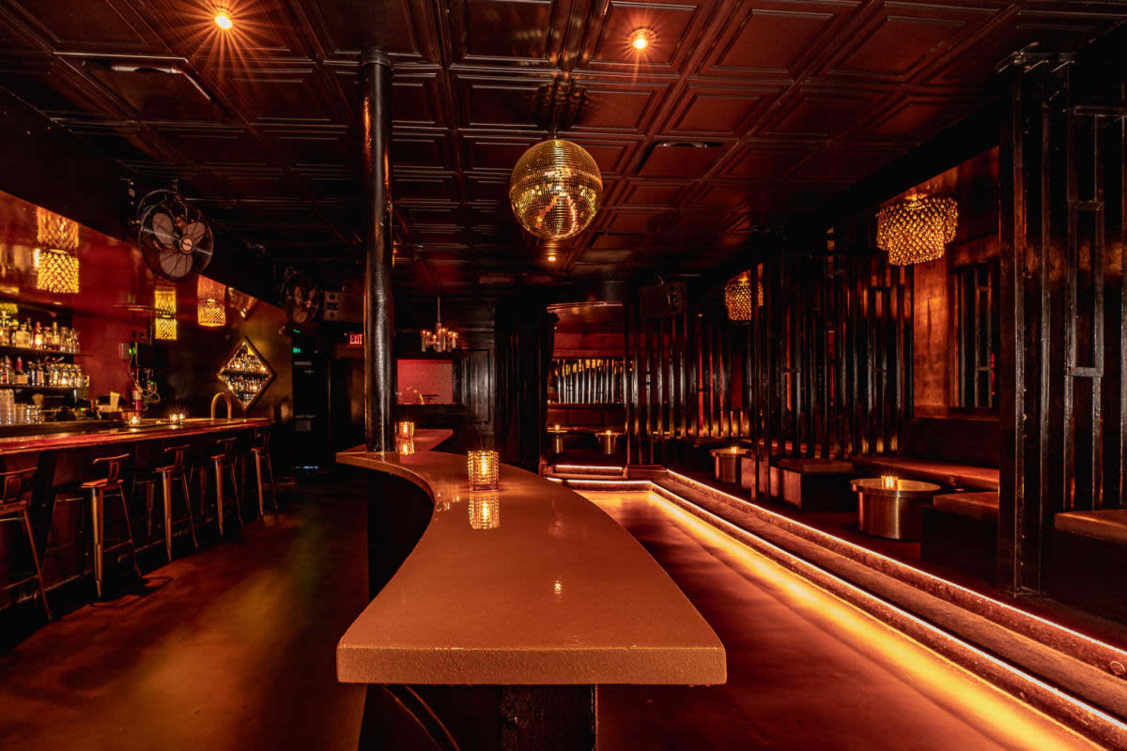 The image depicts a dimly lit bar interior with a curved counter, seating areas along the walls, and decorative lighting fixtures.