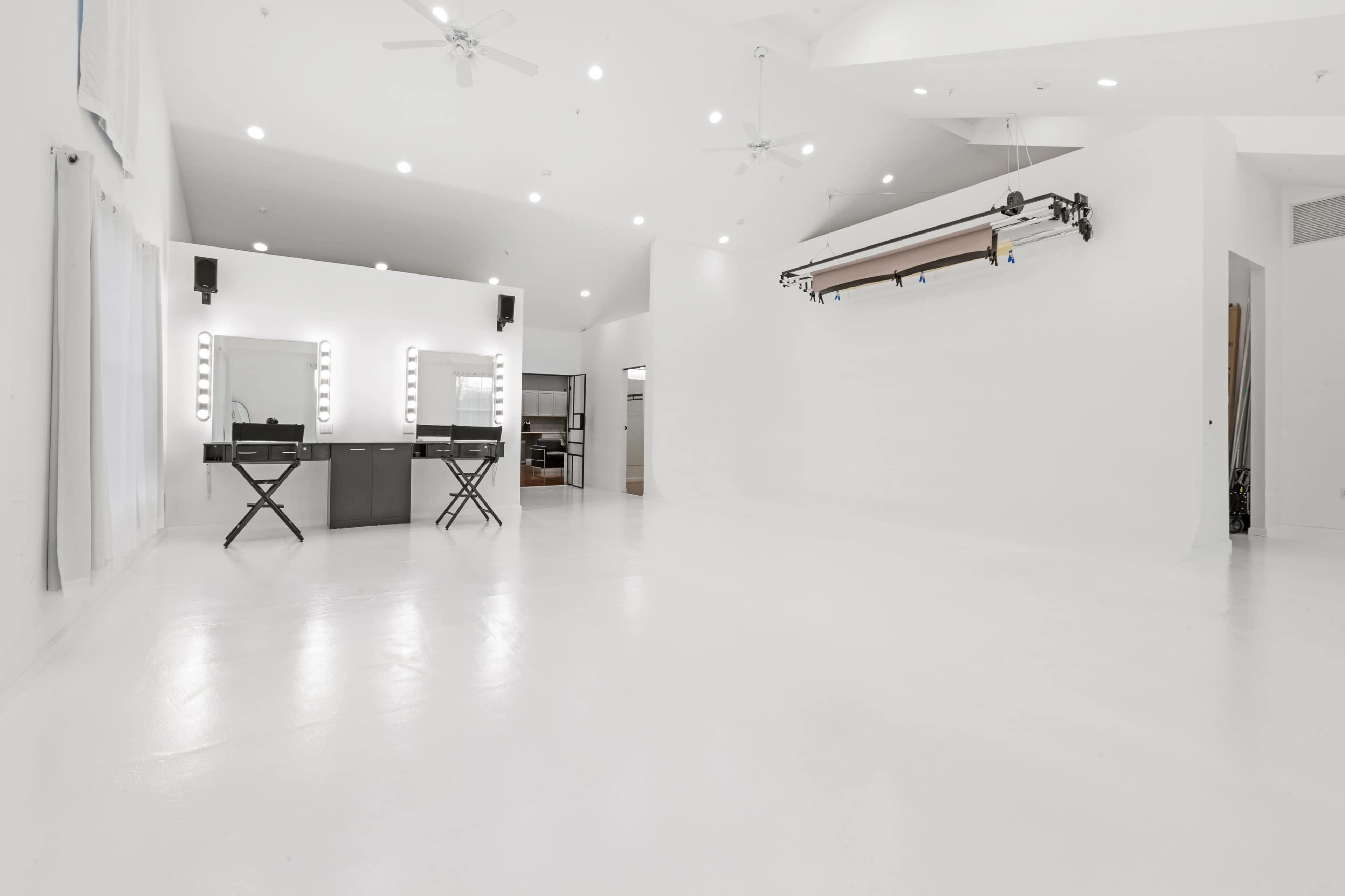 A spacious, white-walled studio featuring two illuminated mirrors and a backdrop system mounted from the ceiling.